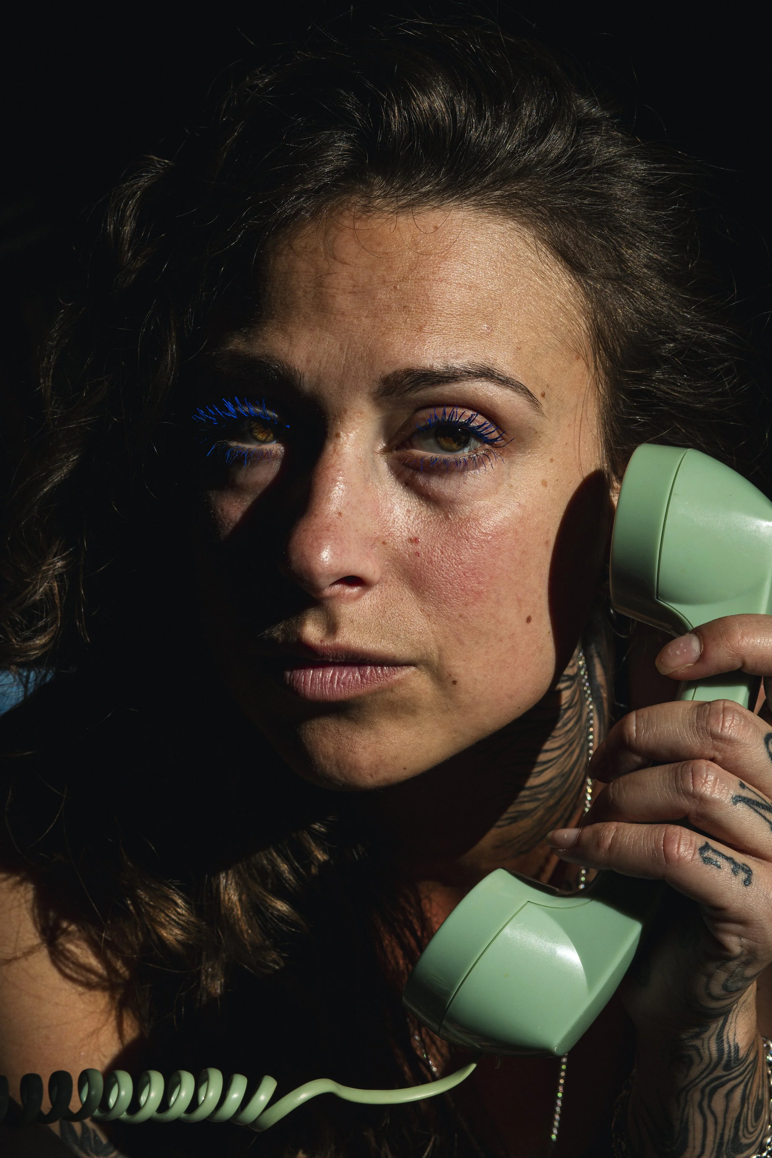 Une femme parle au téléphone avec un appareil vintage de couleur vert pastel, portant du maquillage avec des cils bleus et avez des tatouages sur le bras, dans un environnement sombre.