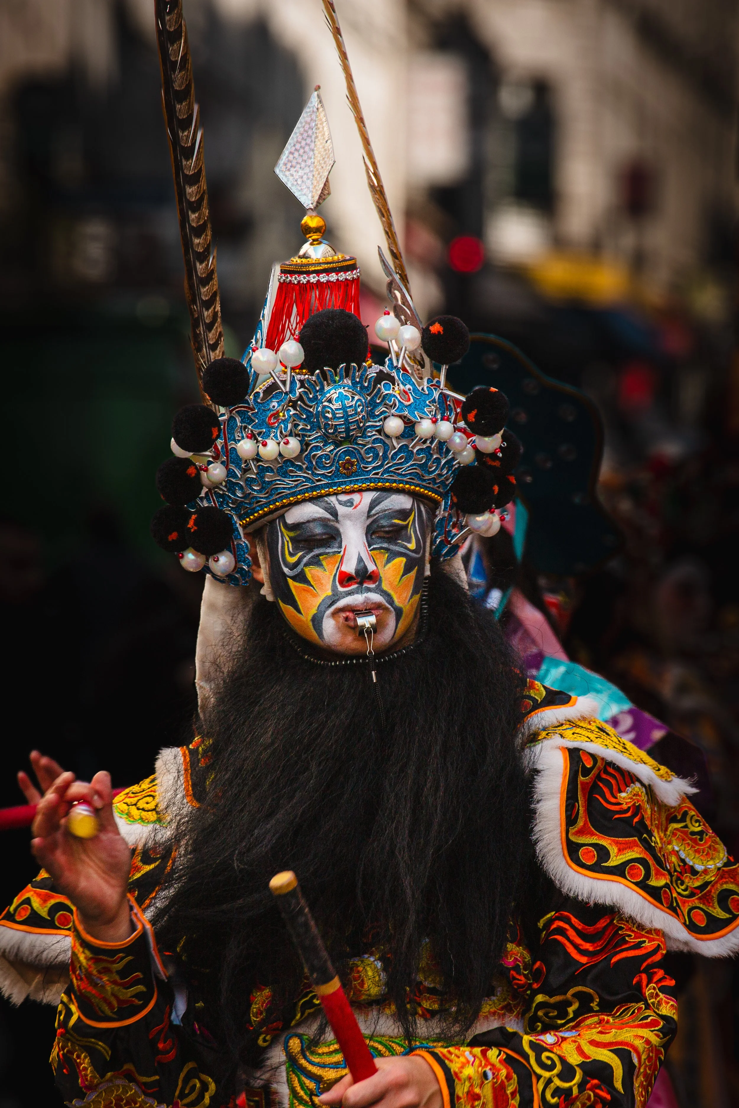 Une personne en costume traditionnel avec un maquillage coloré du visage, portant une coiffe ornée de perles, de plumes et de décorations, dans un contexte probablement festif ou culturel.