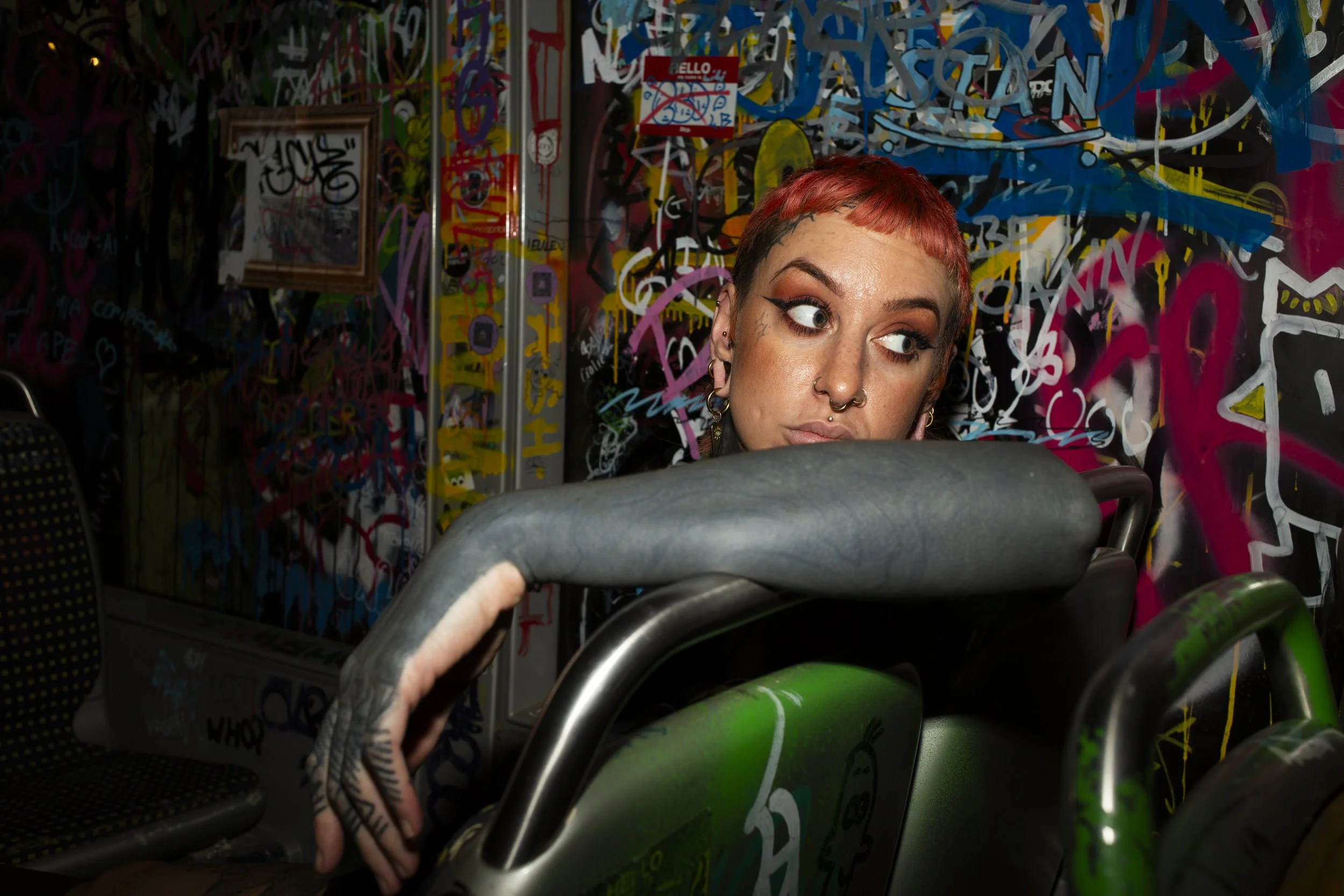 Personne aux cheveux courts roux et tatouages, assise dans un environnement de rue ou d'art urbain avec un mur décoré de graffitis colorés, vue partielle de la tête et de la main tenant un objet, probablement un guidon de vélo.