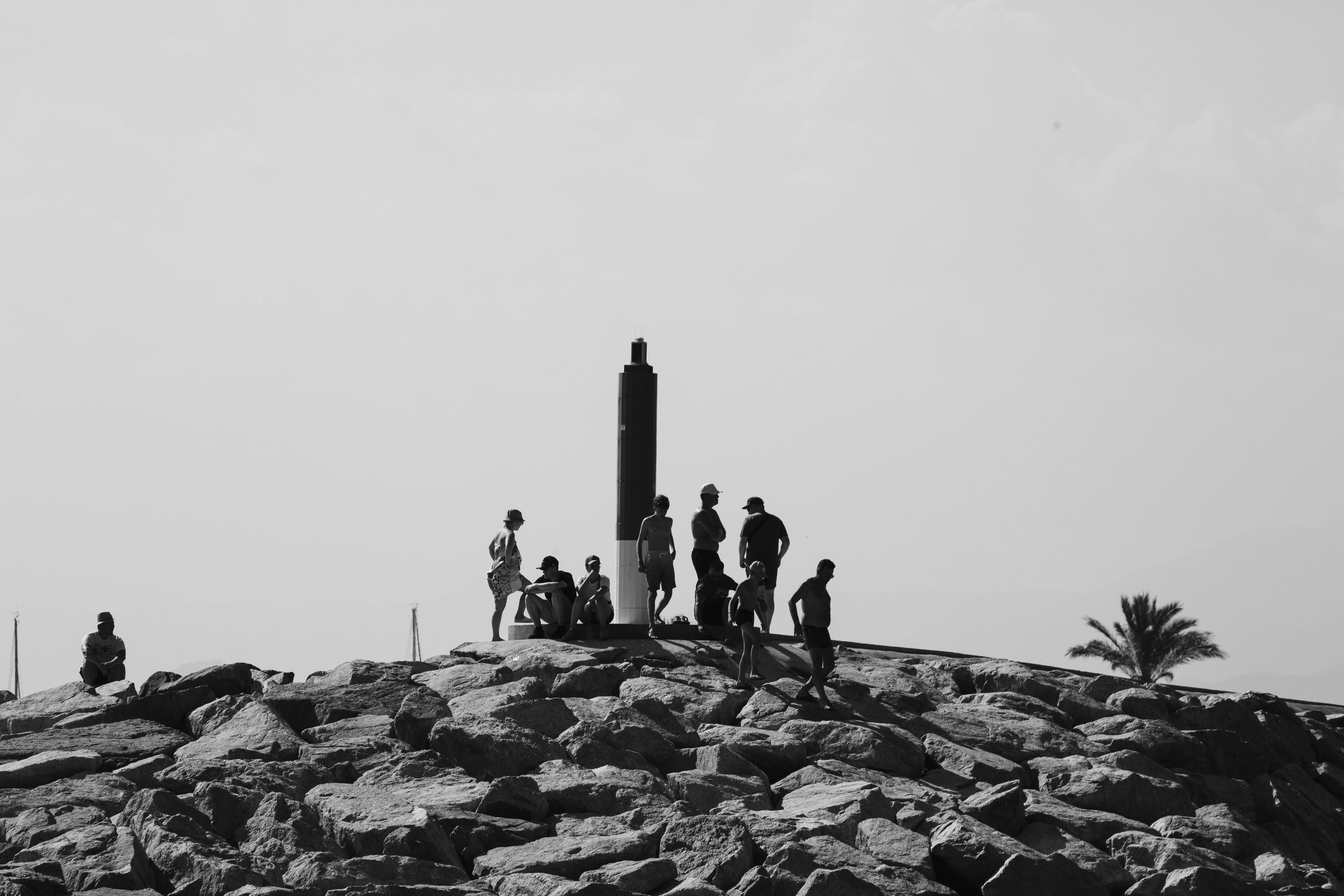 Groupe de personnes assises et debout sur des rochers près d'un monument en forme de tour, avec un arbre à palme en arrière-plan.