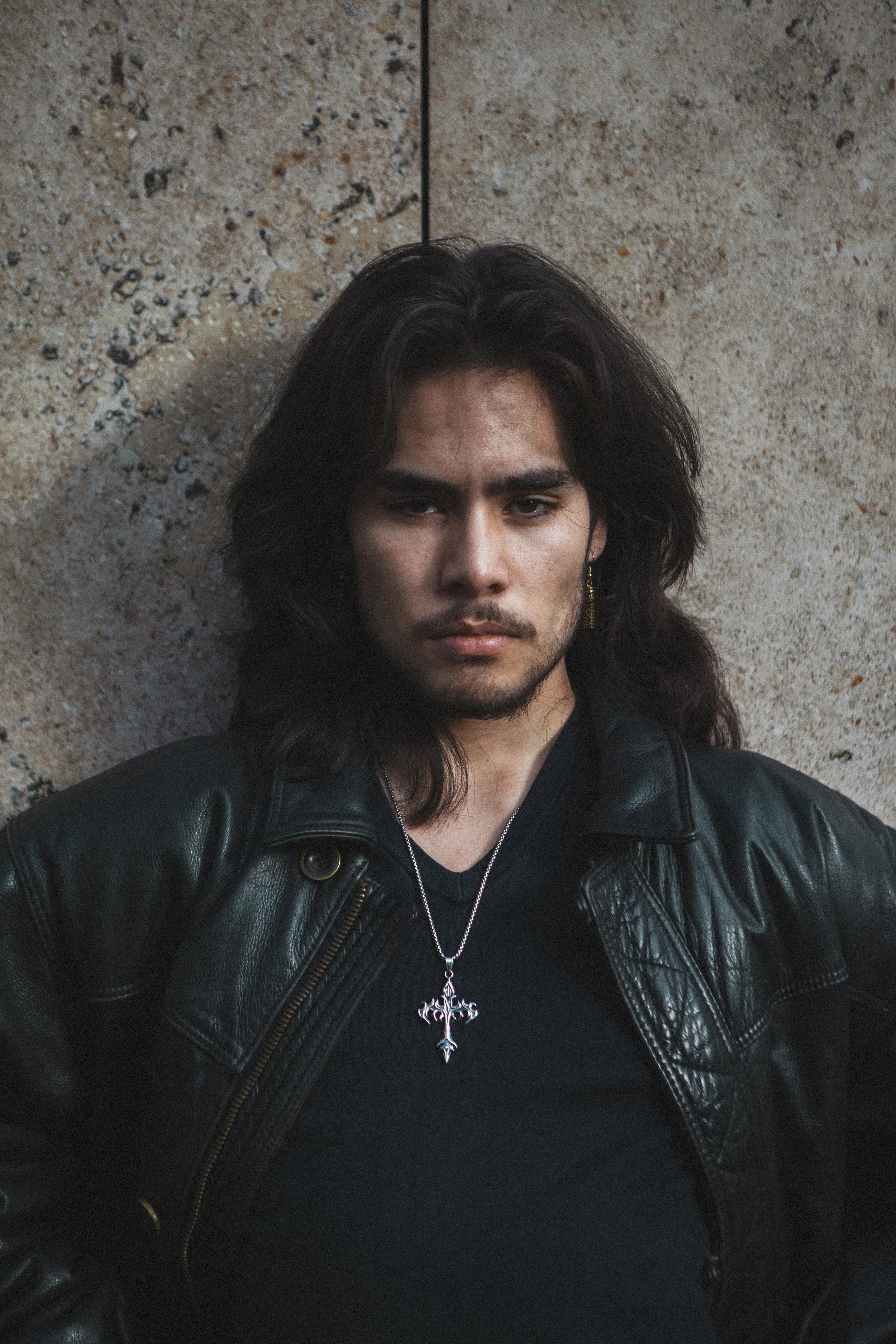 Portrait d'un jeune homme à cheveux longs, portant une veste en cuir noire, une chaîne avec une croix et une oreille à boucle d'oreille, posé contre un mur en béton.