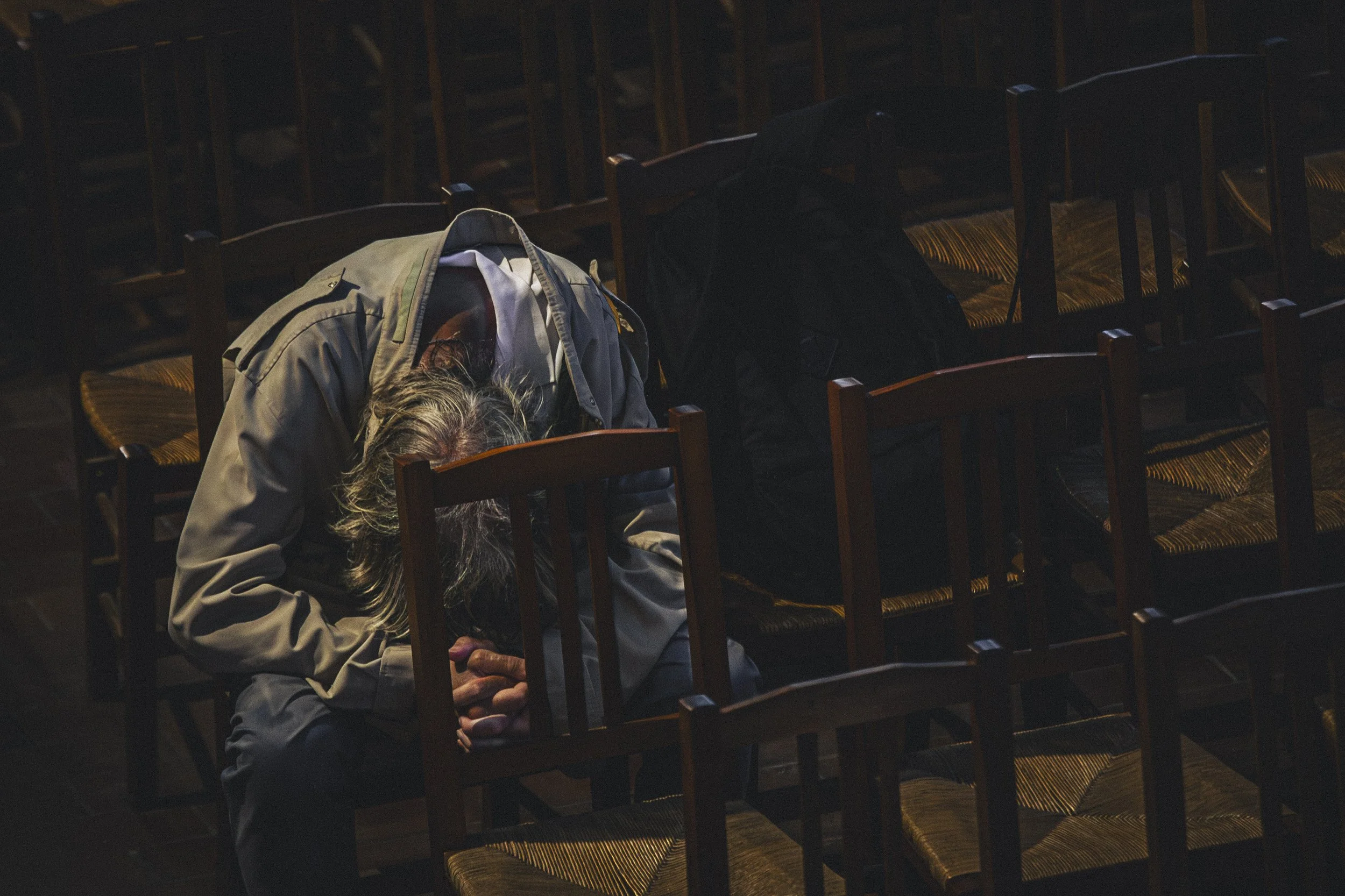 Une personne âgée, probablement un homme, avec des cheveux gris, est assise, les yeux fermés, en prière ou en méditation dans une pièce sombre, entourée de chaises en bois.
