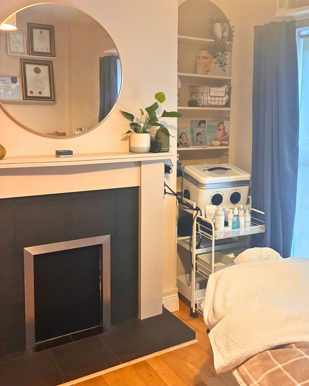 Living room corner with a fireplace, a round mirror above the mantel, a potted plant, and a shelf with skincare products. There is a laundry machine and trolley with bottles beside a window with blue curtains.