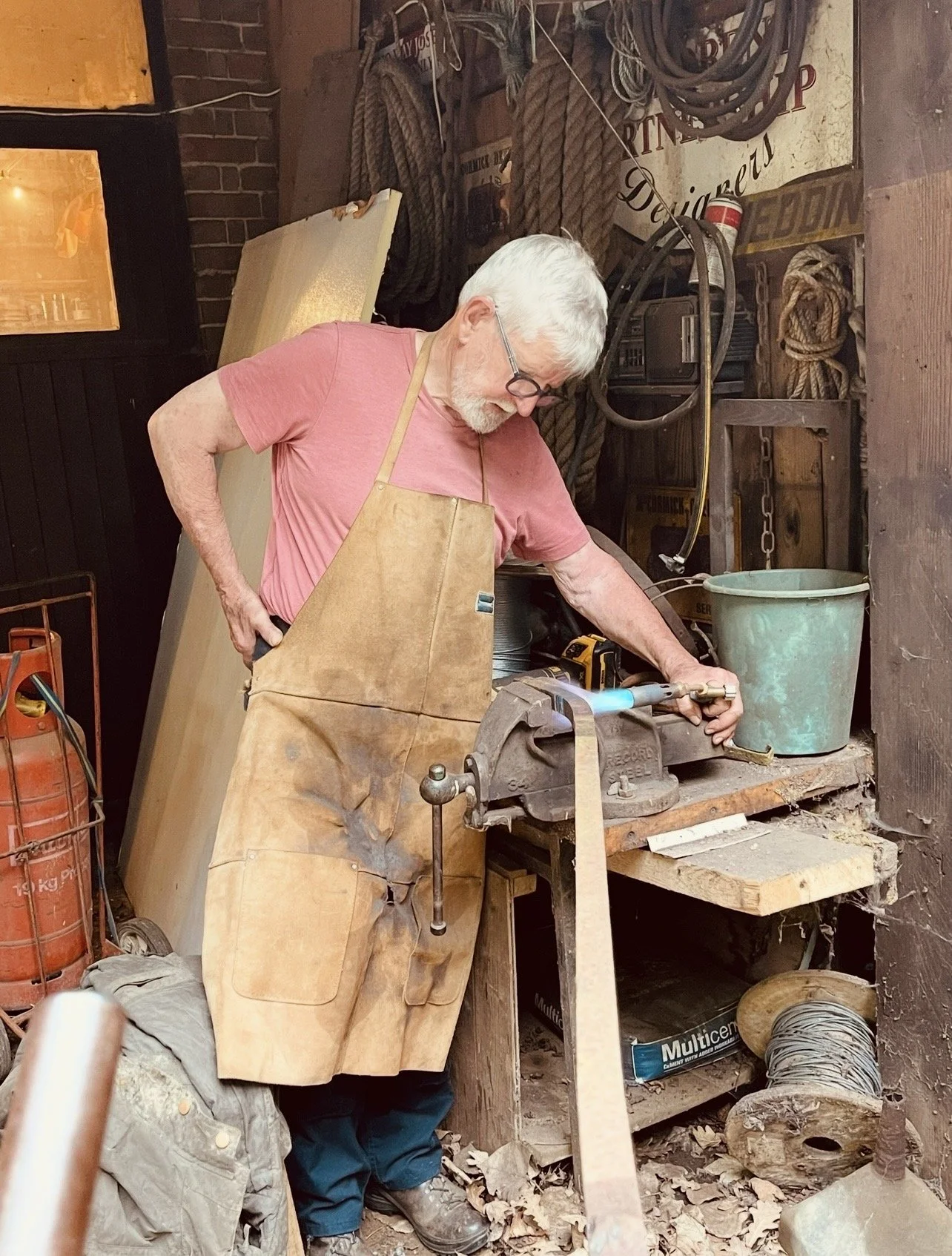 An older man with white hair and glasses wearing a pink shirt and brown apron working with a saw in a woodworking shop. He is cutting a piece of wood secured in a bench vice. The workshop has various ropes, tools, and equipment hanging or sitting on shelves around him.