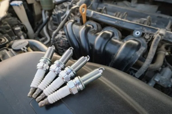 Six spark plugs placed on an engine's surface inside a car engine bay.