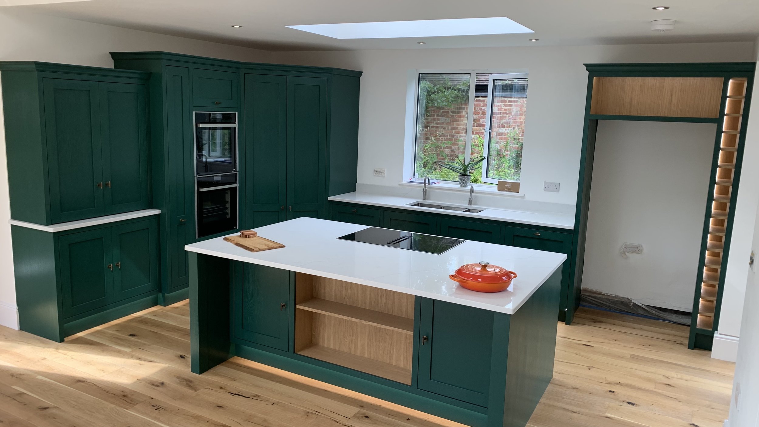 Modern kitchen with teal cabinetry, white countertops, a kitchen island with a wooden base, a built-in oven, a window with a houseplant, and hardwood floors.