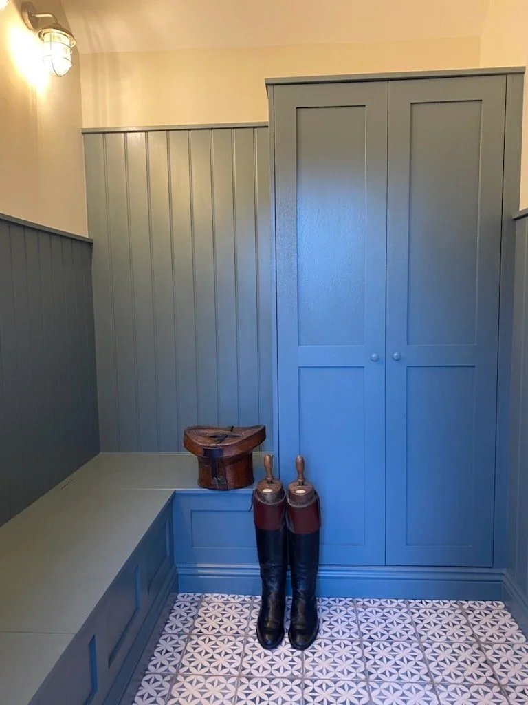 Blue painted built-in cabinet and bench seating corner with a pair of black riding boots and a vintage leather saddle on top of the bench, patterned tile floor, and beige walls with wall-mounted light fixture.