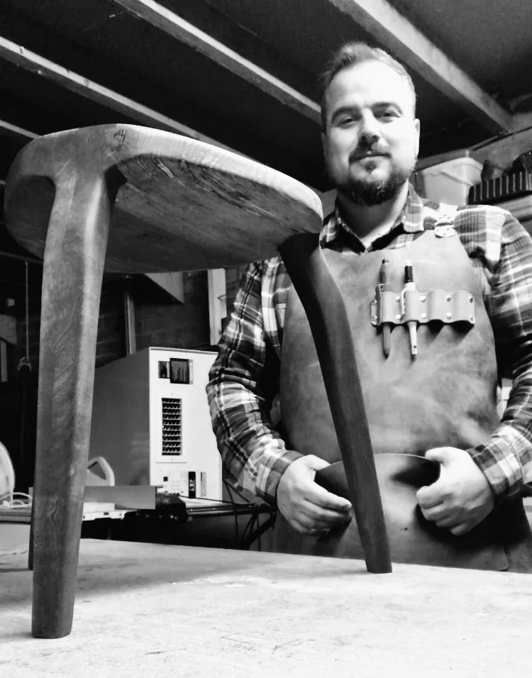 Black and white photo of a man in a woodworking shop, smiling, wearing a flannel shirt and apron, holding a chisel. In front of him is a wooden stool he likely crafted.