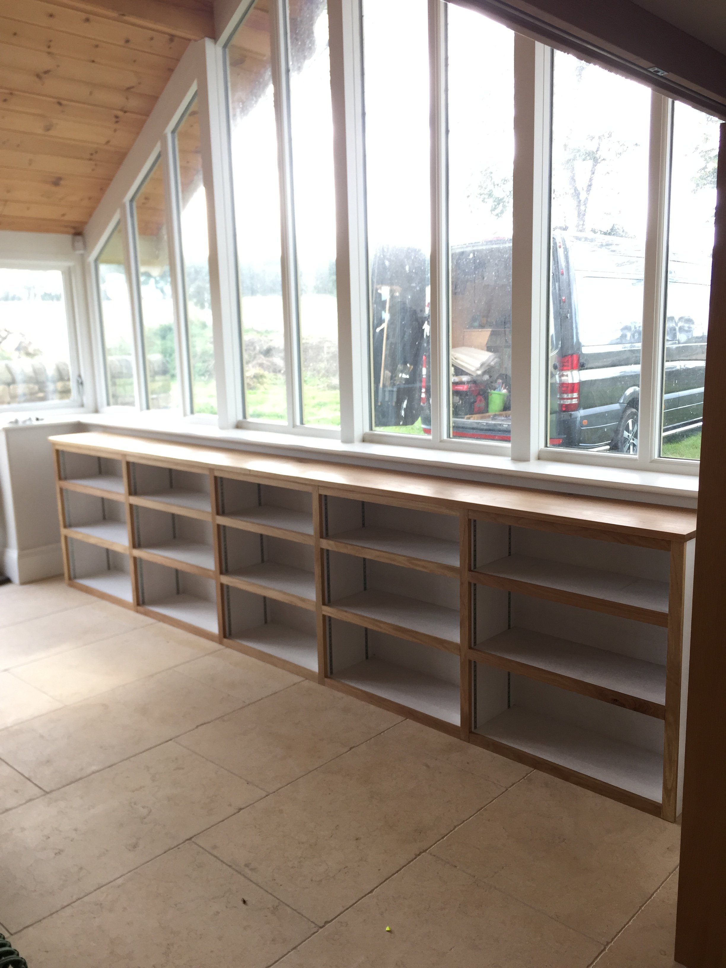 unfinished wooden bookshelf near a large window in a room with beige tiled floor and wood-paneled ceiling.