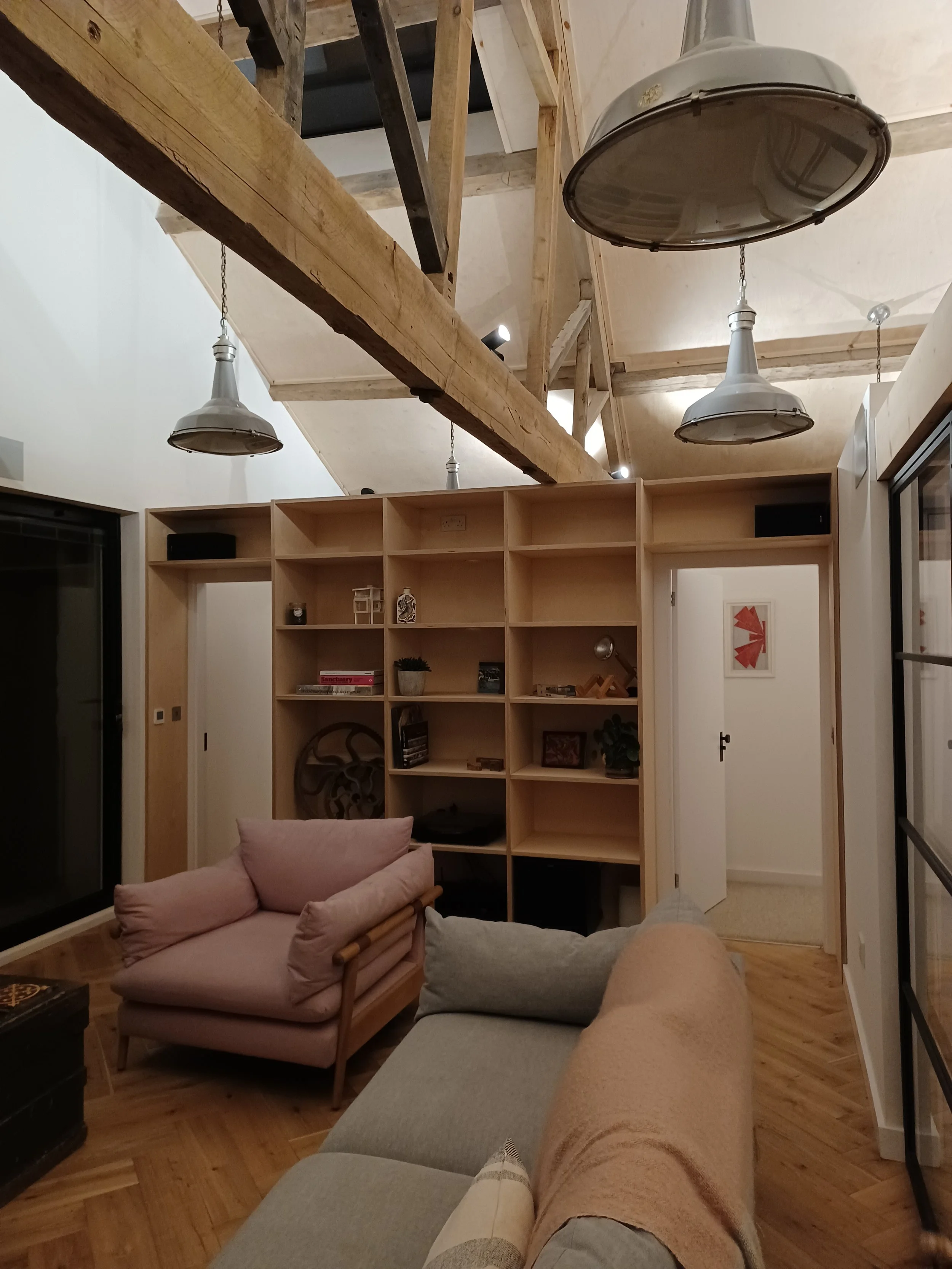Living room with beige sofa, pink armchair, open wooden shelving unit, exposed wooden beams on ceiling, hanging industrial-style lights, window with black frame, and a framed abstract art piece on the wall.