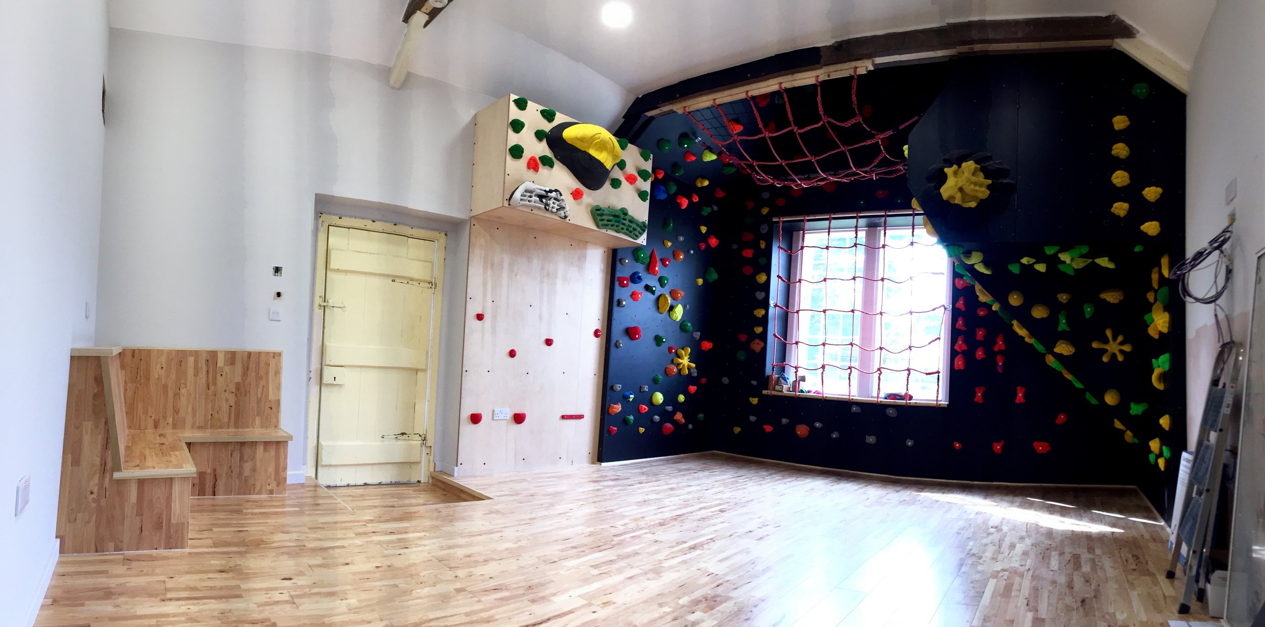 An indoor bouldering gym with colorful climbing holds on the walls and a large climbing net hanging from the ceiling.