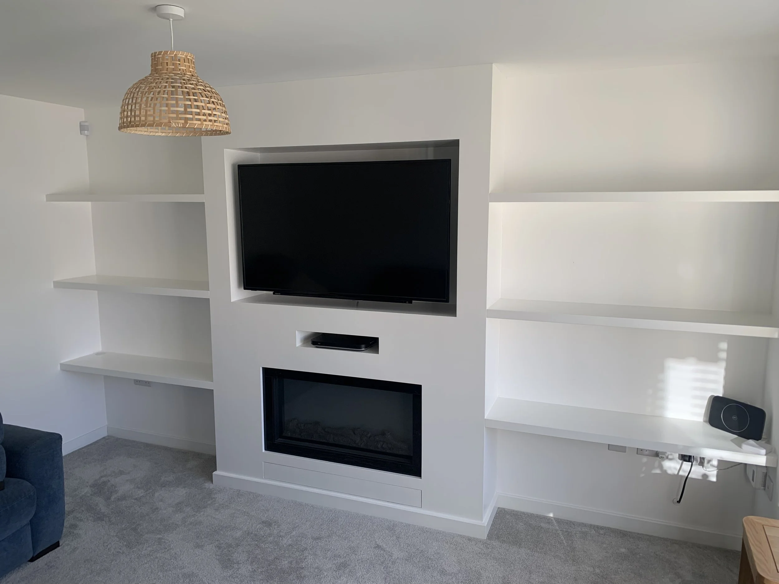 Living room with white built-in shelving unit and a black flat-screen TV mounted in the center. A television remote is on a white shelf. There is a gray carpeted floor and part of a blue sofa is visible on the left.