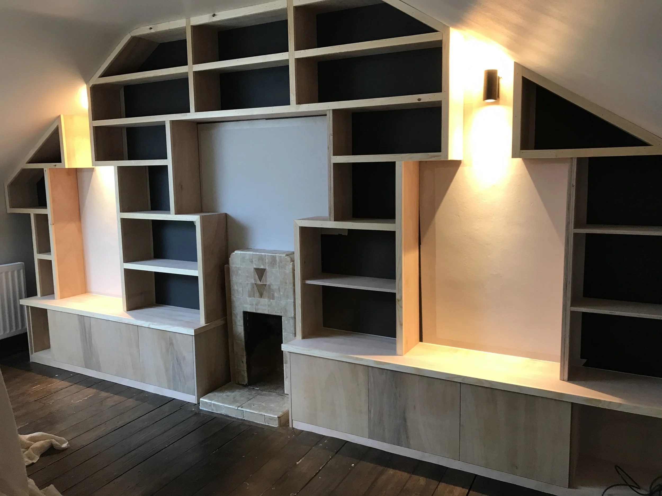 An unfinished wooden shelving unit with multiple compartments, built into an angled wall in a room with dark wood flooring and light-colored walls.