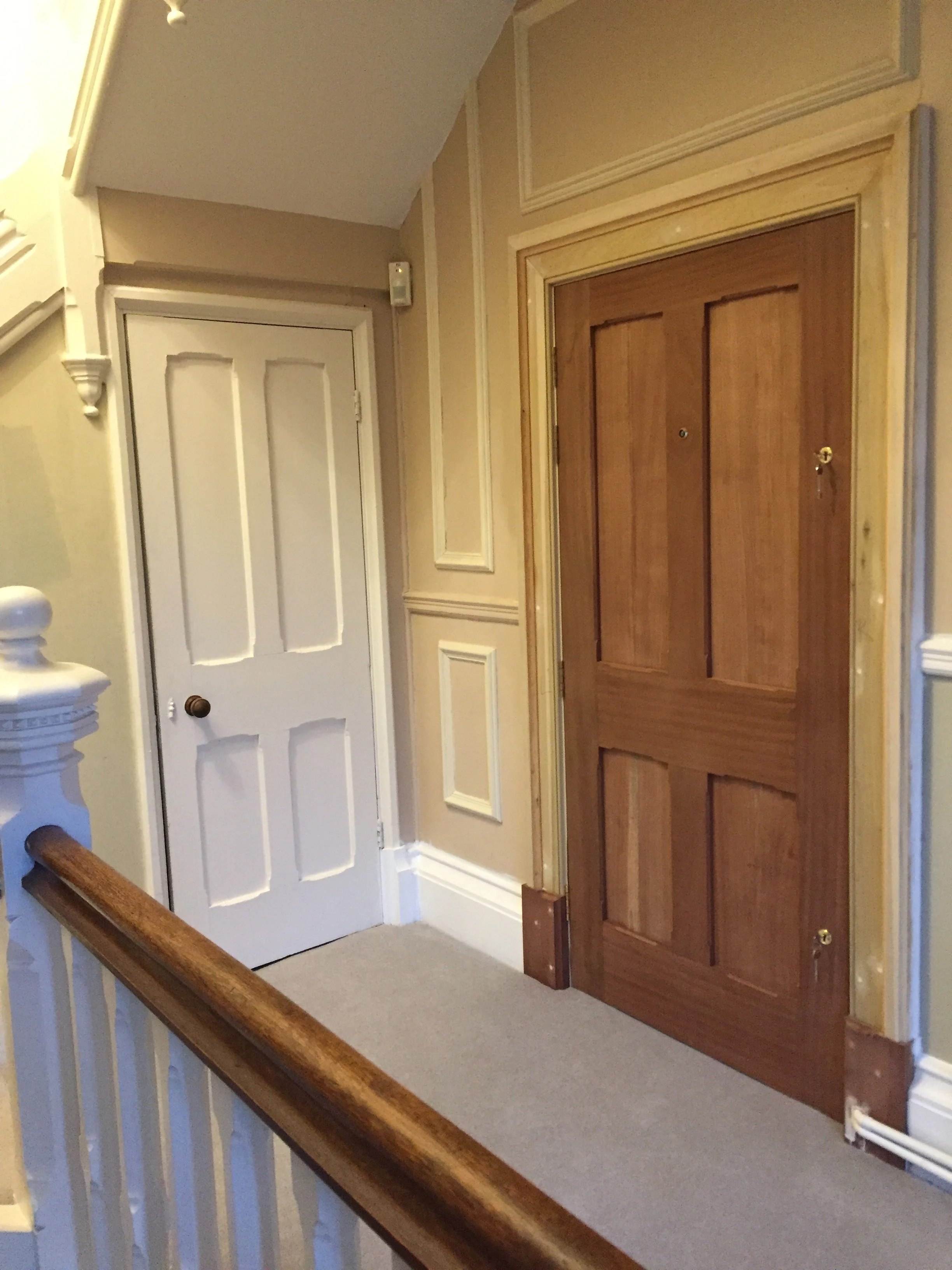 Interior view of a hallway with a white front door and a wooden elevator door, beige walls with decorative trim, and a wooden handrail.