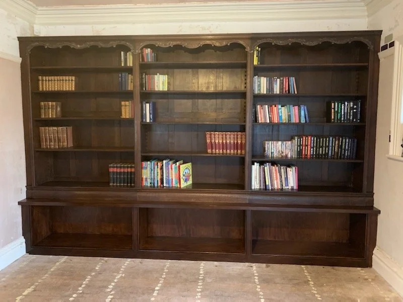Dark wooden bookshelf with closed cabinets at the bottom, containing scattered books with some groups of books on the shelves, positioned against a white wall next to a window.
