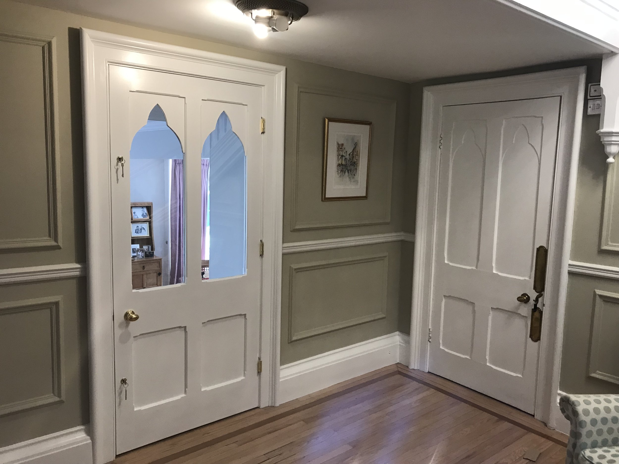 A hallway with light olive green walls, decorative white wainscoting, and two white doors with arched glass panels. The door on the left has a small picture frame with multiple photos, while the door on the right has a matching decorative molding. A 