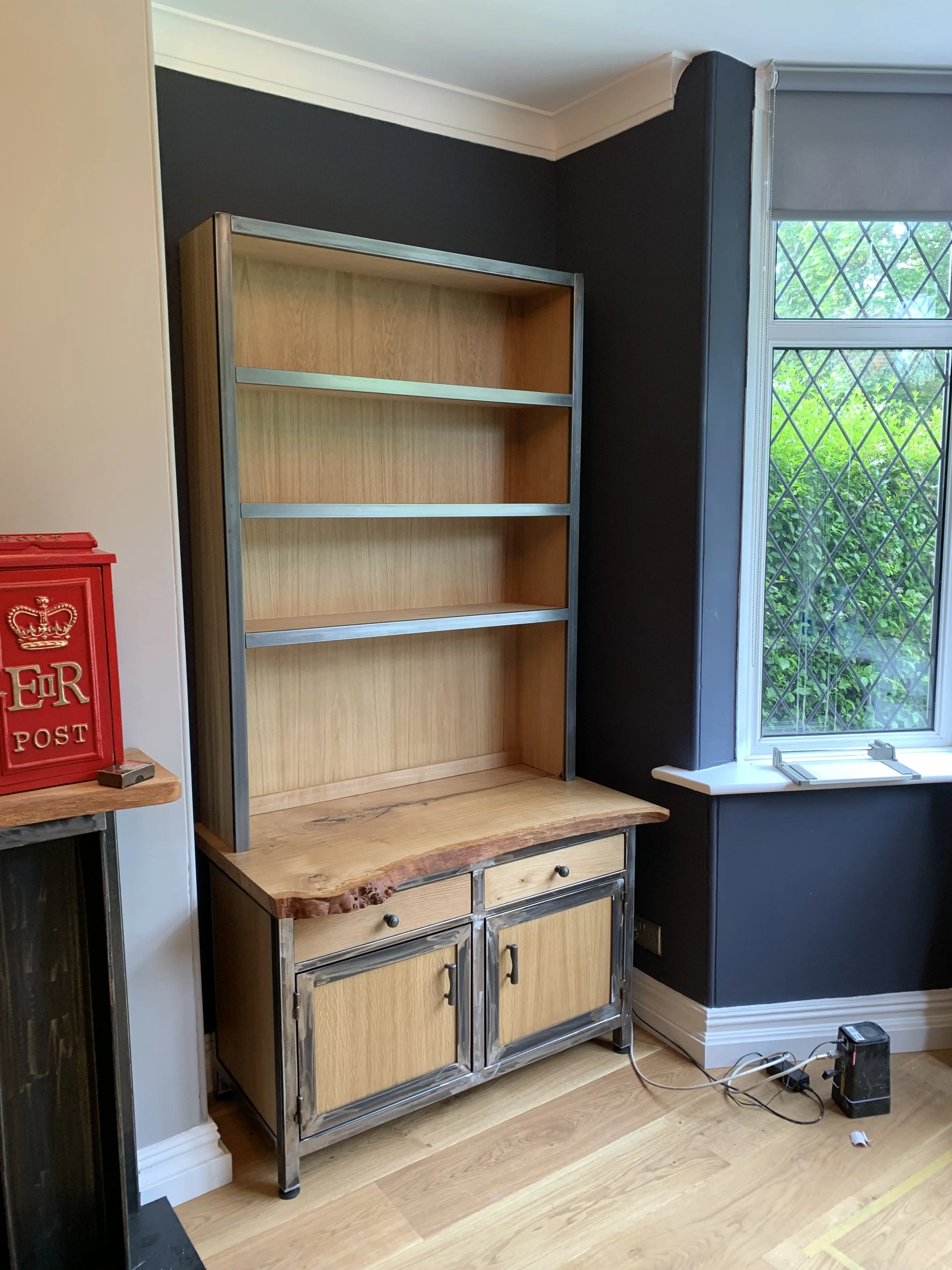 A partially assembled wooden kitchen cabinet with a metal frame, placed against a black wall next to a window with diamond-shaped grid pattern.