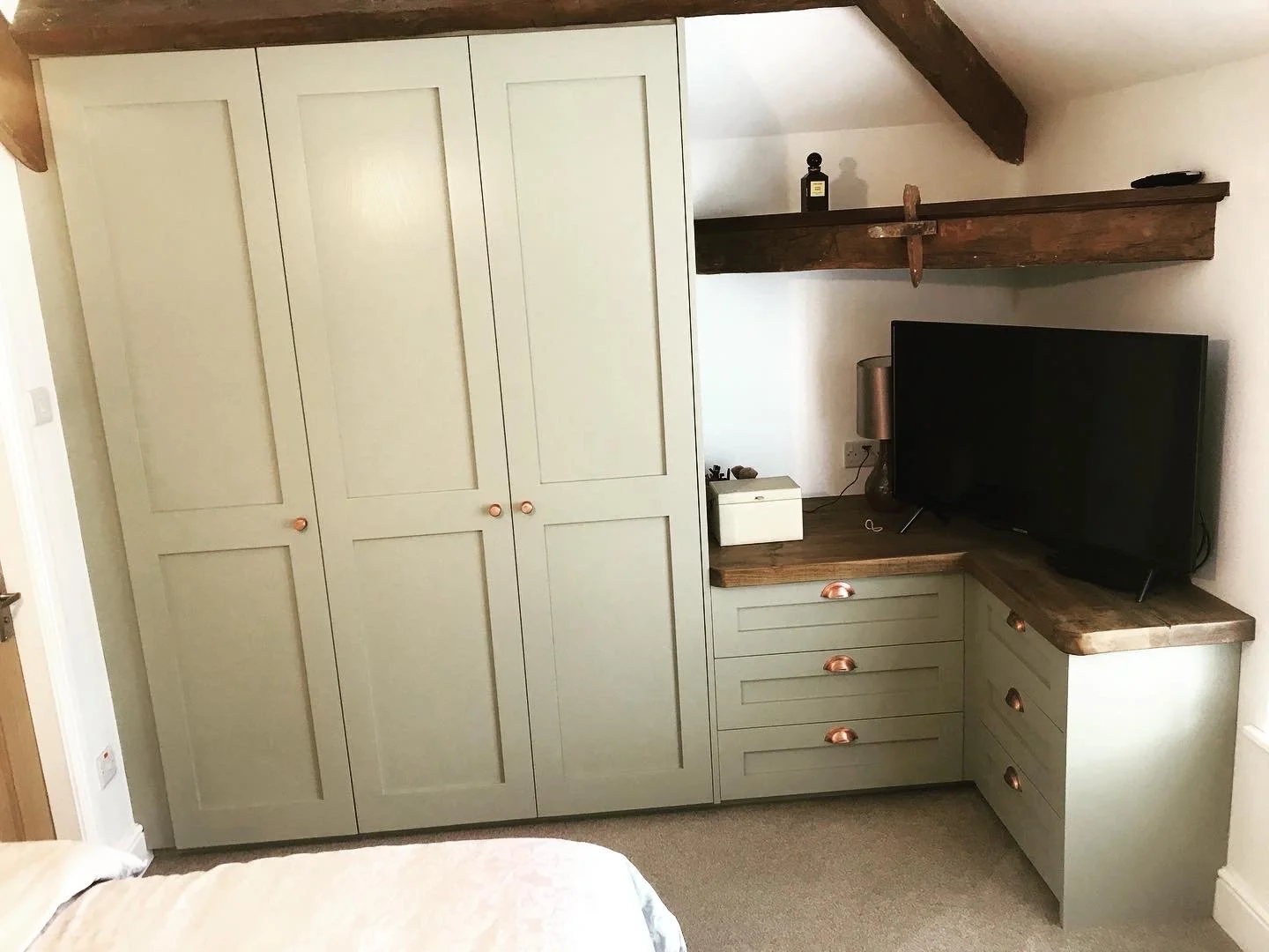 A corner of a room featuring a built-in wardrobe with light green doors and wooden handles, a wooden countertop against the wall with a flat-screen TV, a silver lamp, a white box, and a wooden shelf above holding decorative items including a dark bot