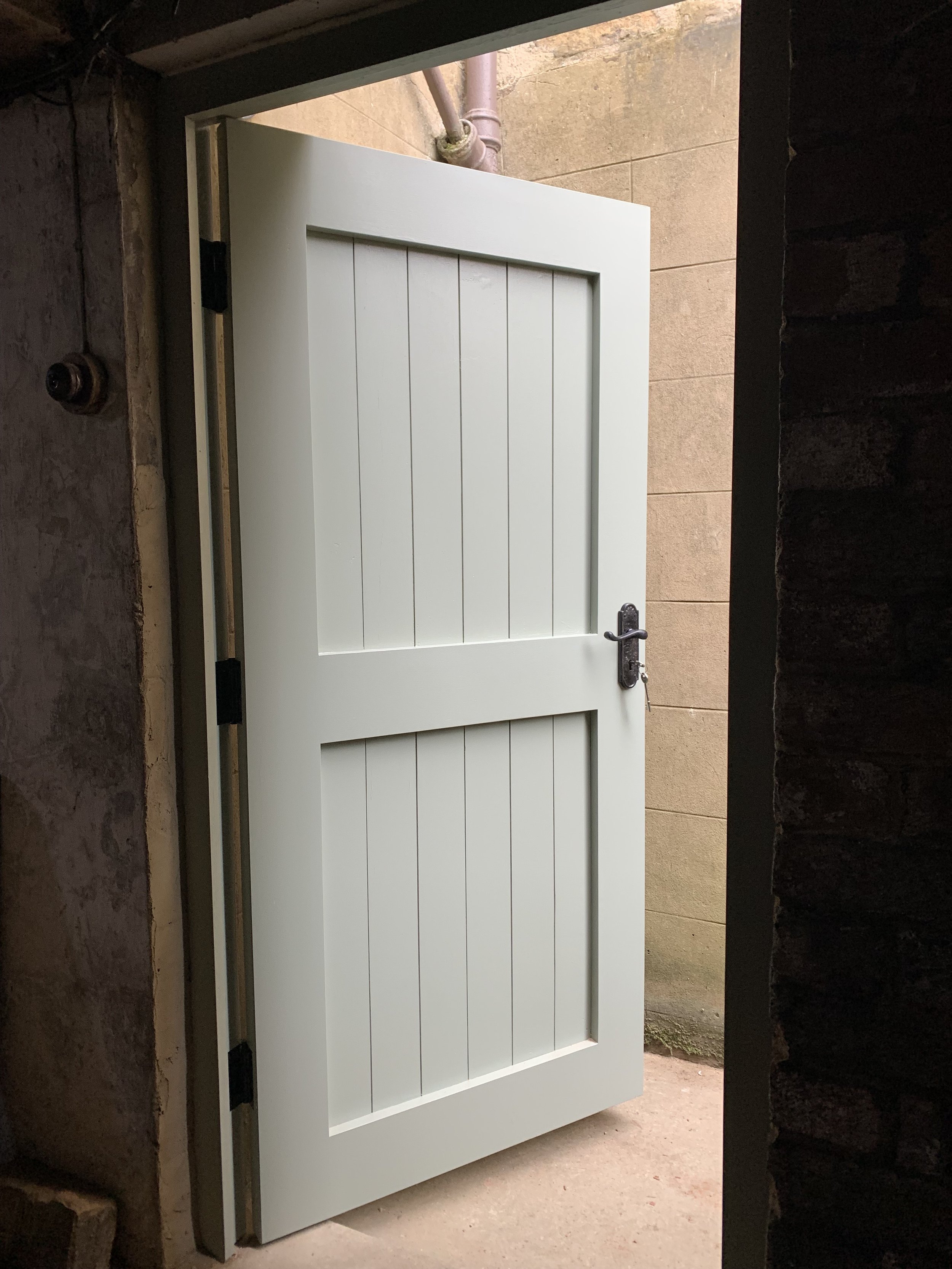 A white door with horizontal panels, slightly open, showing a brown brick wall and a pink pipe outside. The door has a black handle and is mounted in a dark hallway or room.