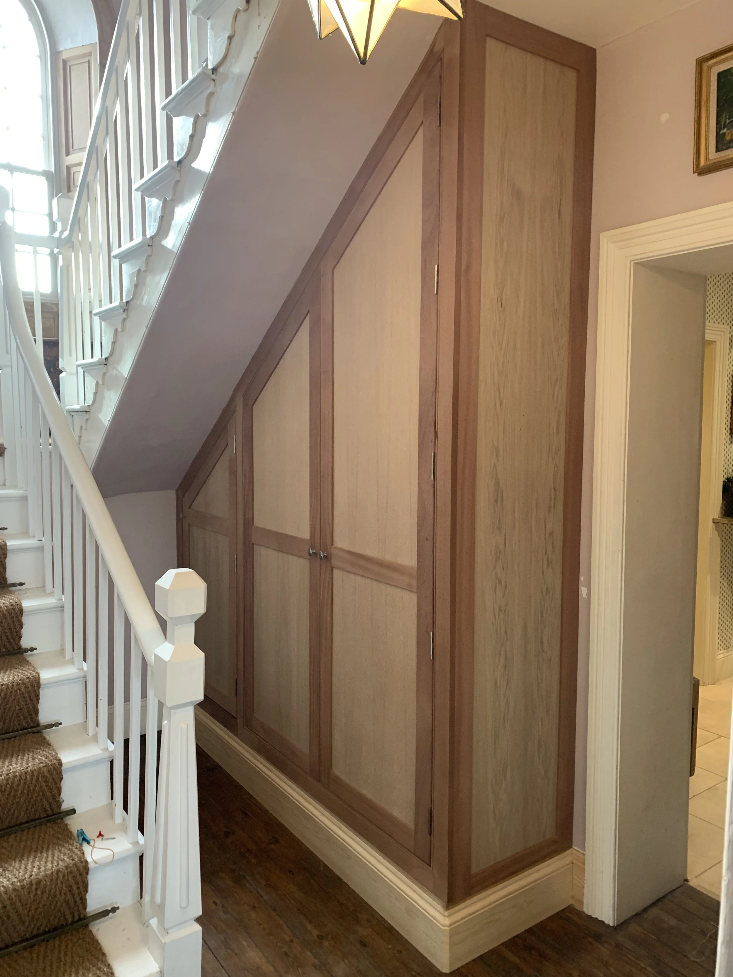 A staircase with carpeted steps and a white railing, leading up to a second floor, situated next to a built-in wooden cabinet with doors, in a house with beige walls and hardwood flooring.