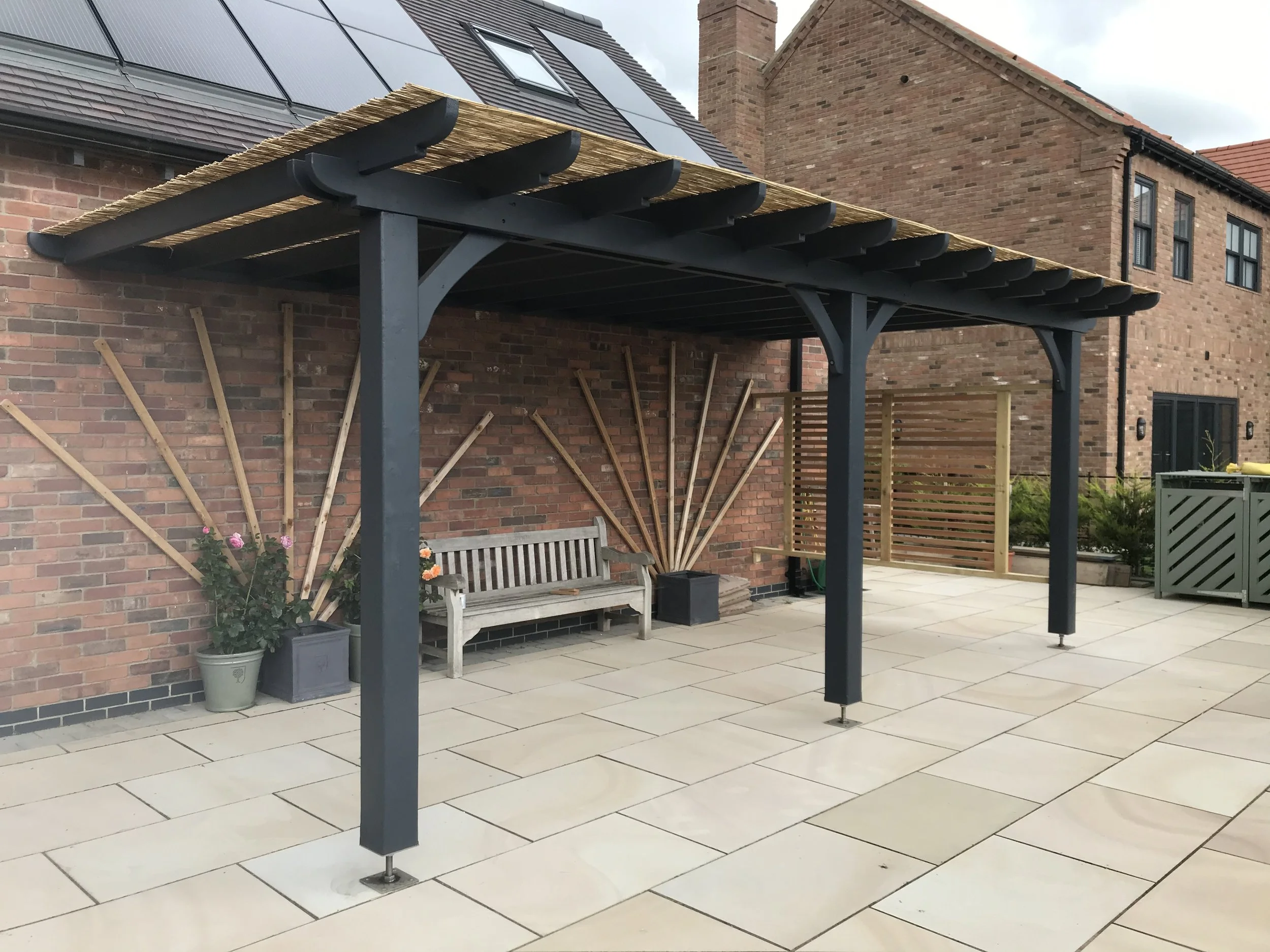 Backyard patio with a black pergola structure, a wooden bench, potted plants, a brick wall, and neighboring brick houses.
