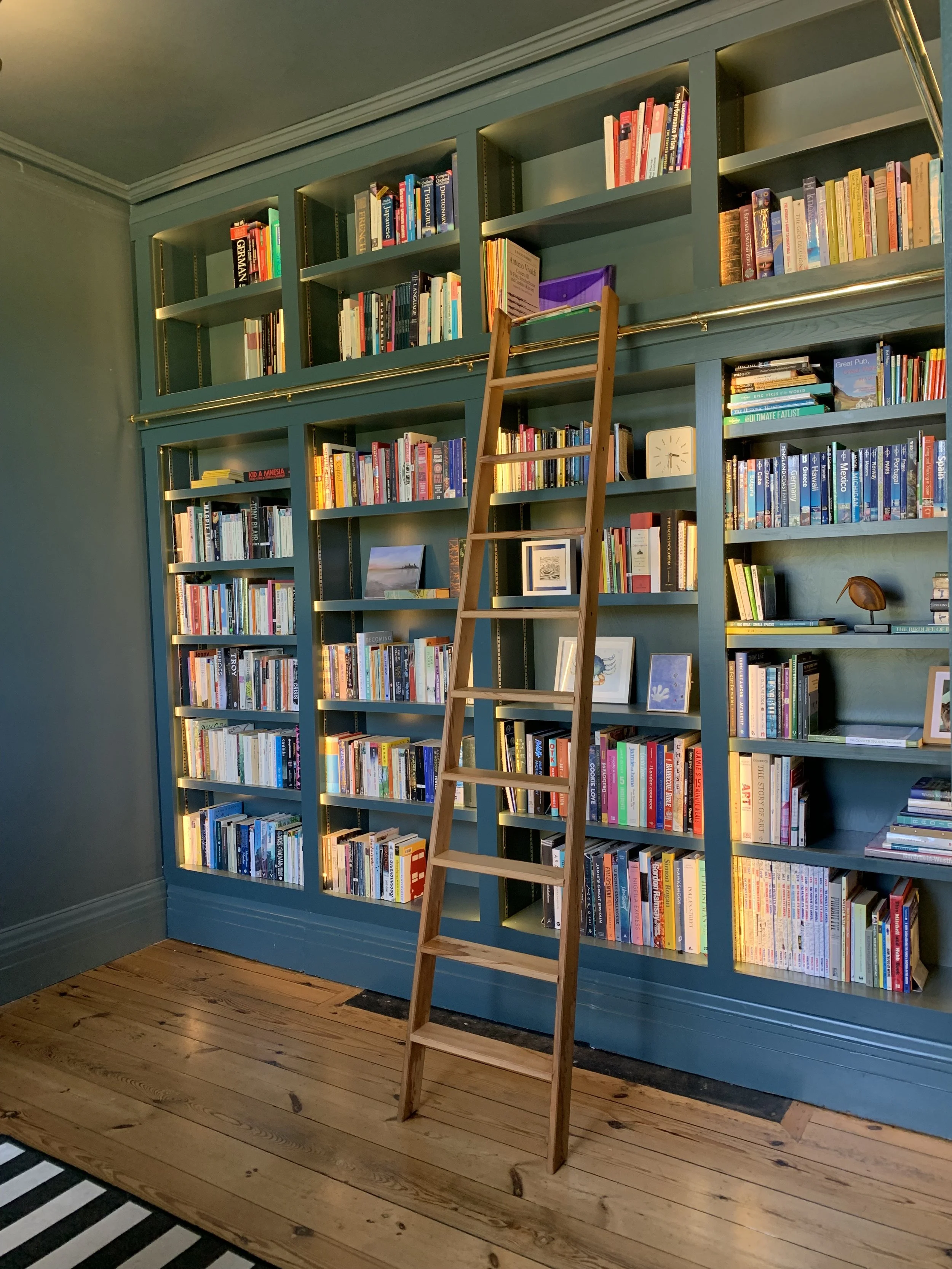 A tall, green bookshelf filled with books against a green wall in a wooden-floored room. A small wooden ladder leans against the bookshelf.