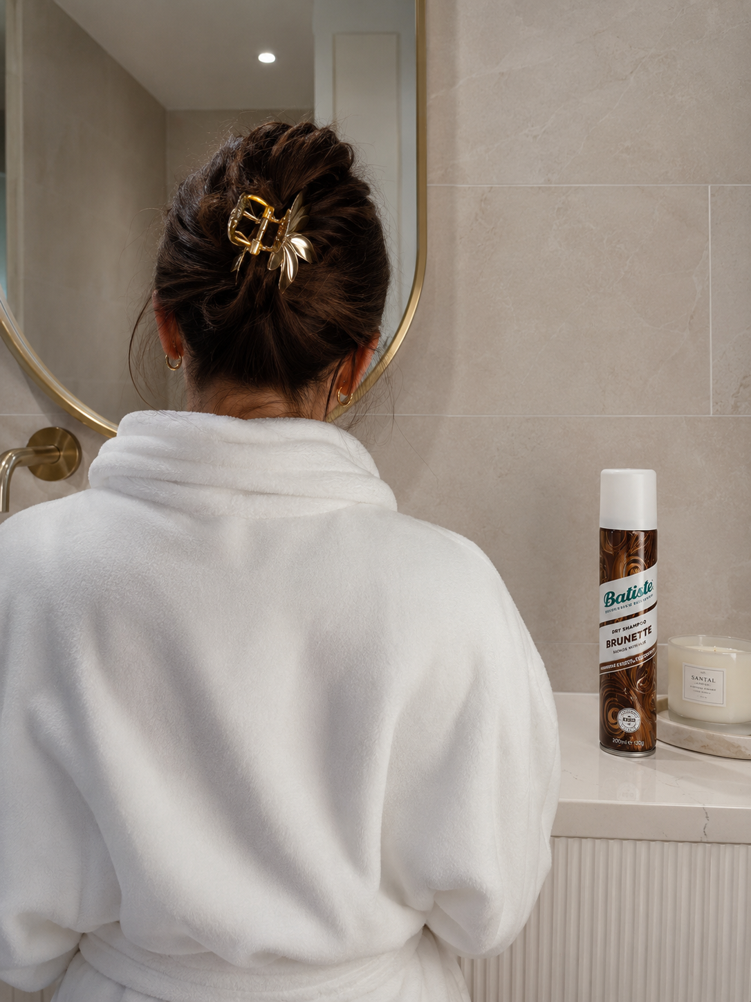 Woman in a white robe with a gold hair clip beside Batiste brunette dry shampoo and a scented candle in a bathroom
