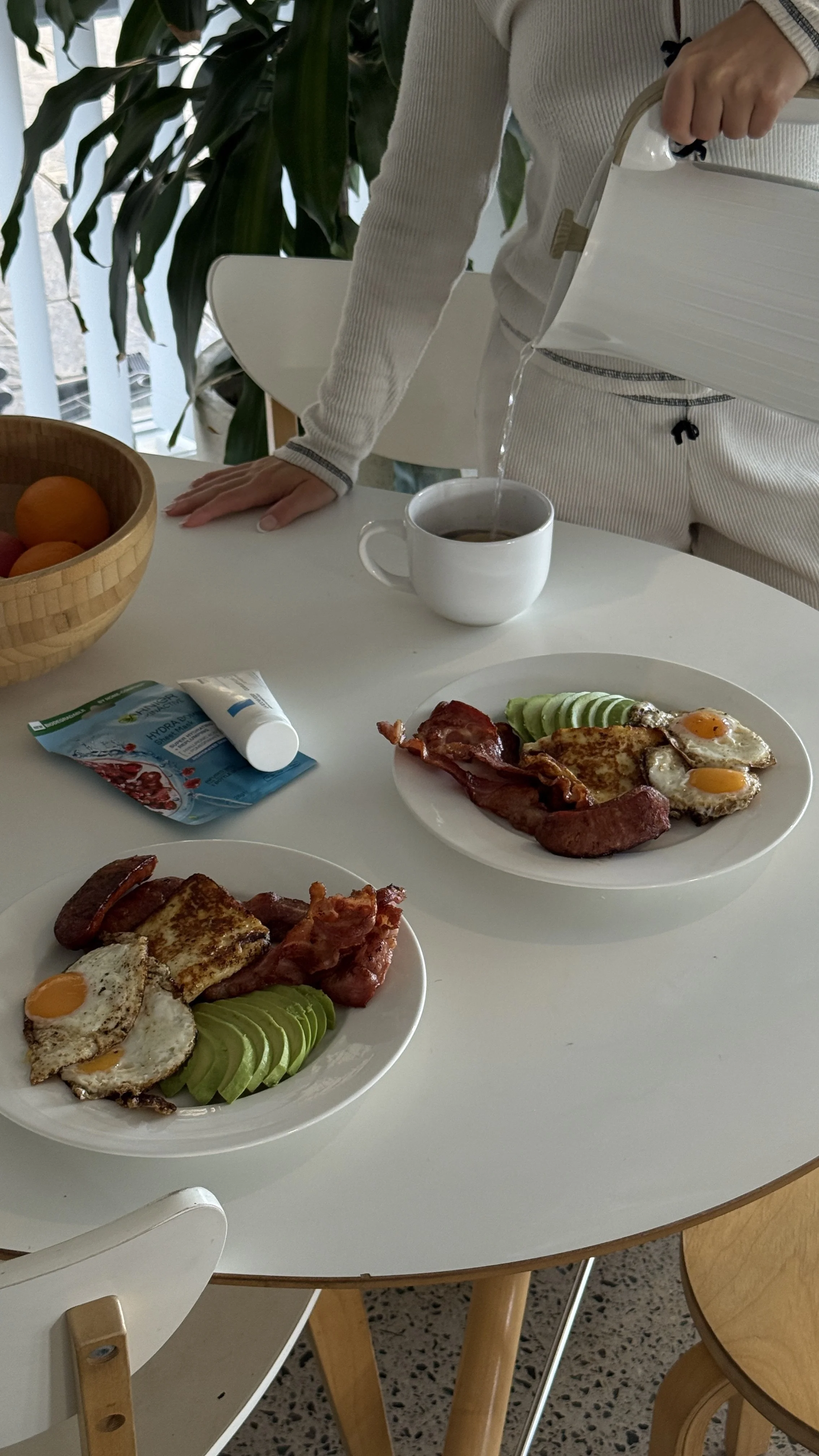 Person pouring tea into a white cup beside plates of fried eggs, bacon, sausage, avocado, and toast at a breakfast table