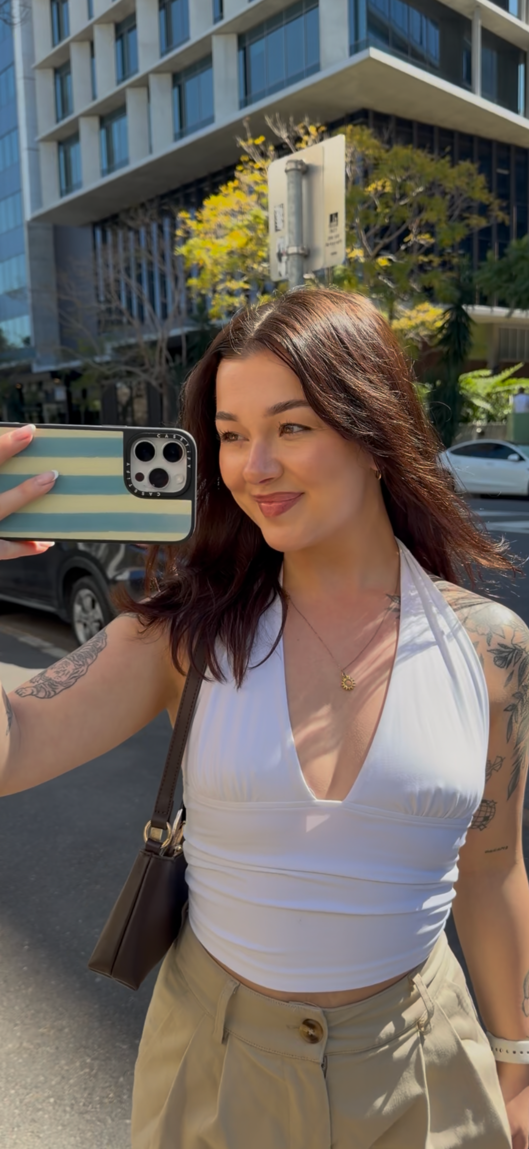 Woman taking a street-style selfie in a white top and beige pants outside a modern building