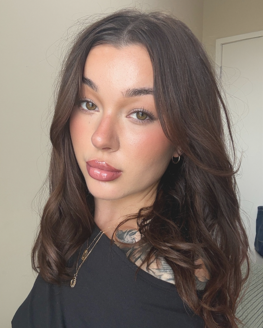 Close-up portrait of a woman with long brown hair, glossy lips, hoop earrings, and a visible chest tattoo wearing a black top