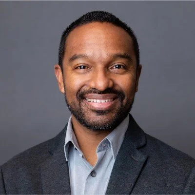 Professional headshot of a Brian Gomez in a gray suit with a light blue shirt, smiling, against a gray background.