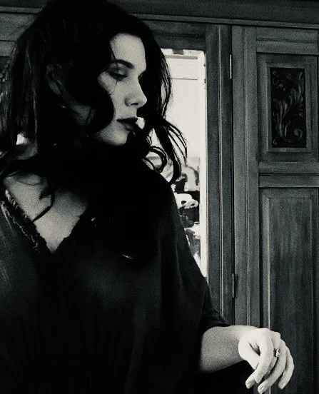 Black-and-white portrait of a woman with long dark hair standing beside a paneled wooden door