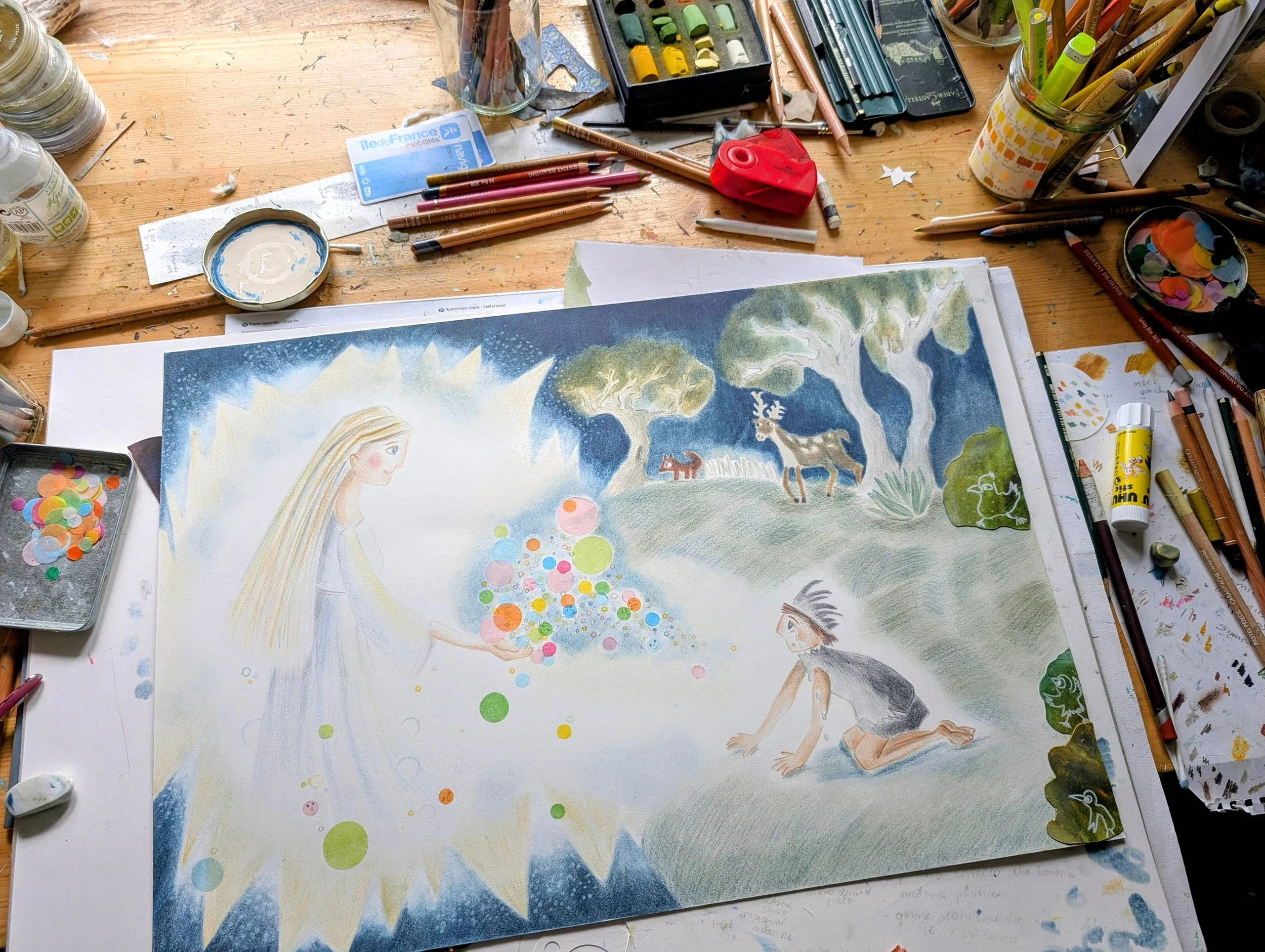 Artist's workspace with a partially completed watercolor painting of a girl, trees, animals, and colorful circles, surrounded by art supplies including colored pencils, paint, and brushes on a wooden table.
