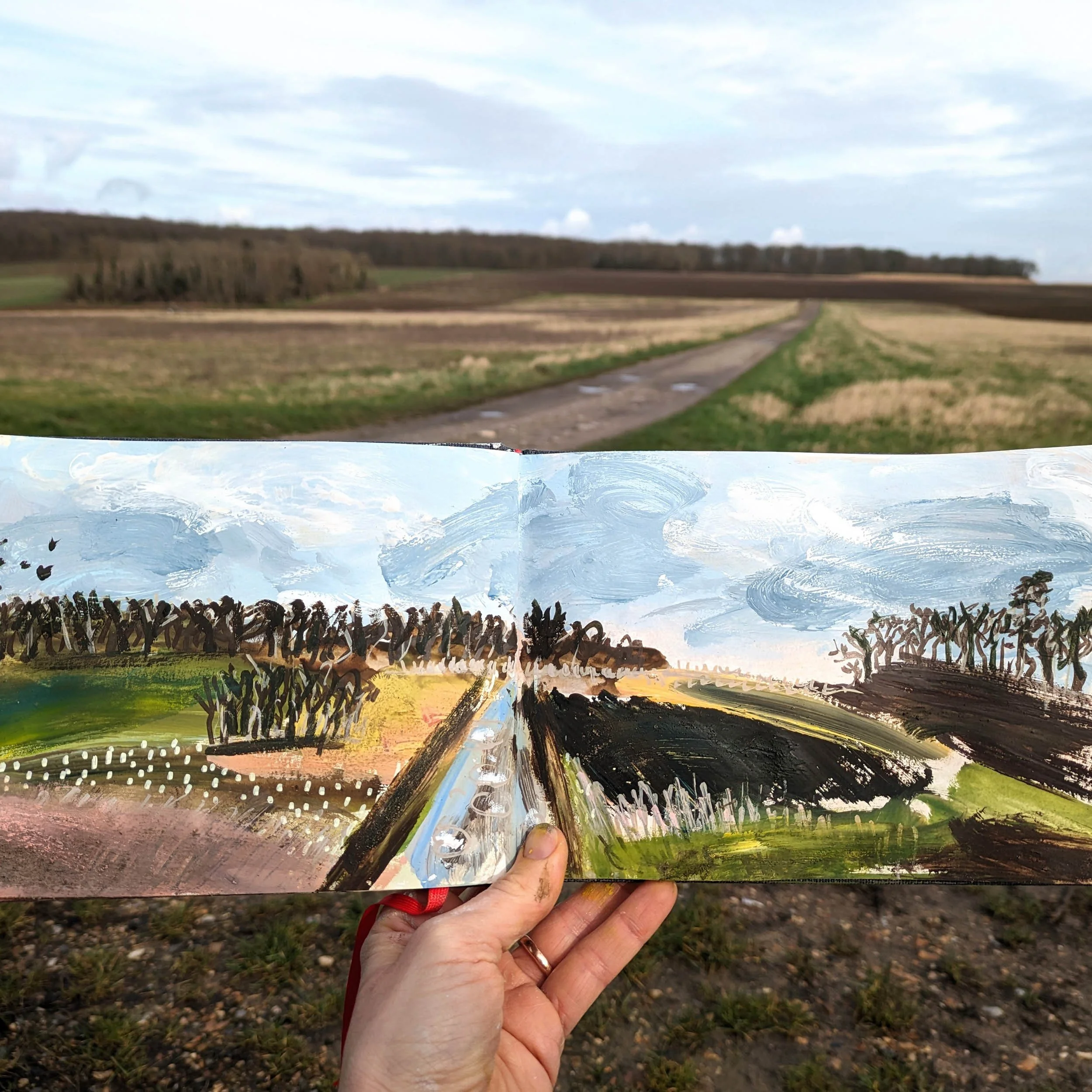A person holding a colorful painting of a landscape scene with fields, trees, a farm, and a sky, against an outdoor background of grass and rocks, in the Yvelines region near Paris.