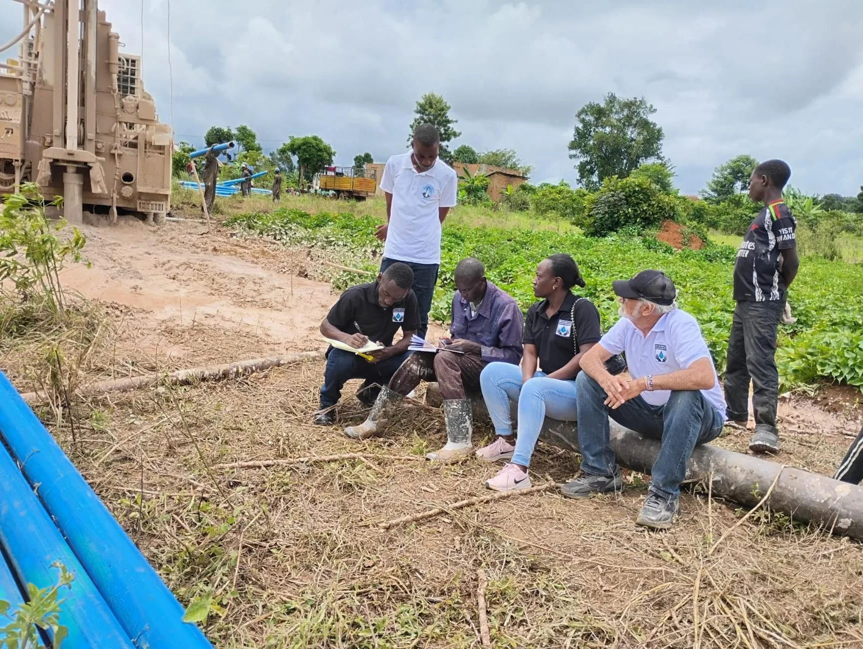 Group of five people outdoors in a field, sitting and standing near a log, with plants and trees around, some individuals taking notes while others observe, and a large construction or drilling machine in the background.