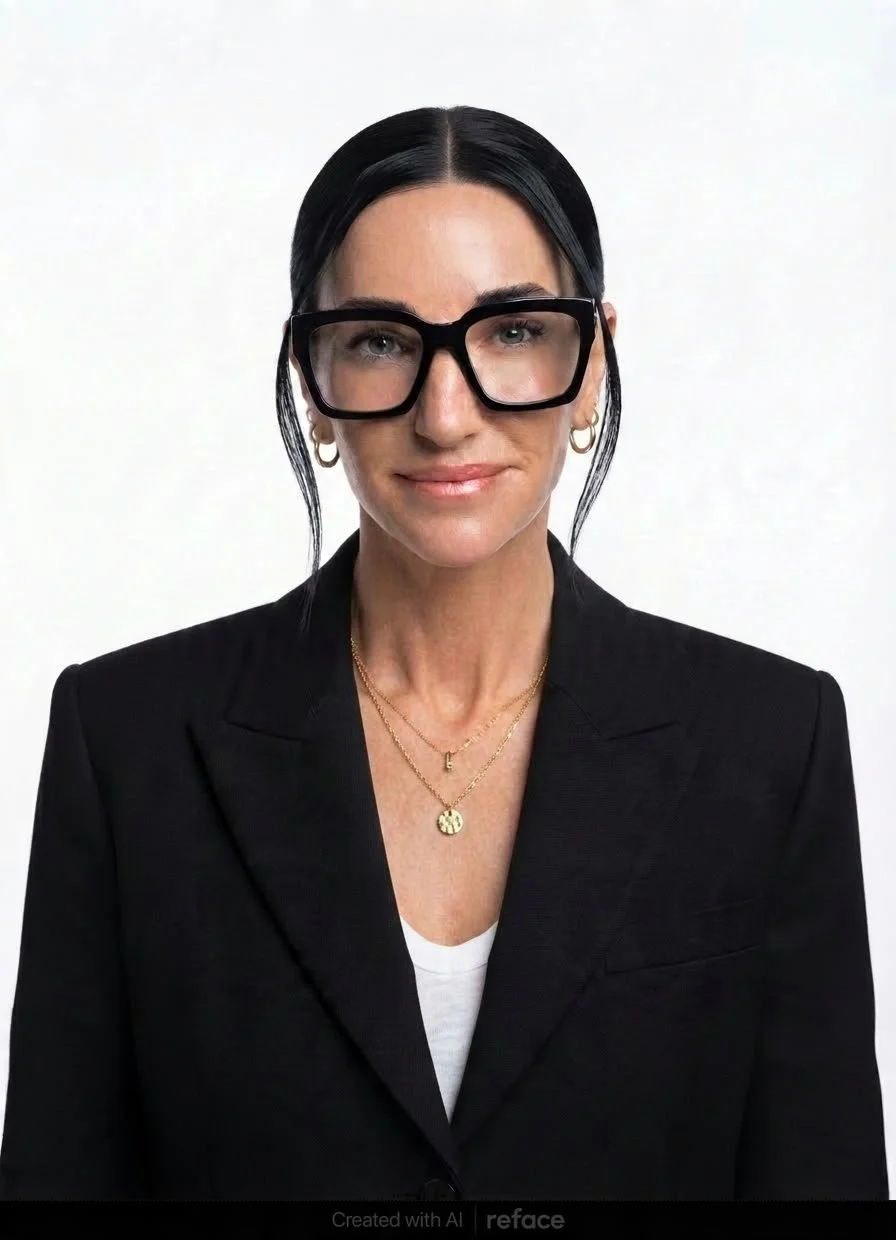 A woman with black hair, glasses, and gold jewelry, wearing a black blazer, smiling at the camera against a plain white background.