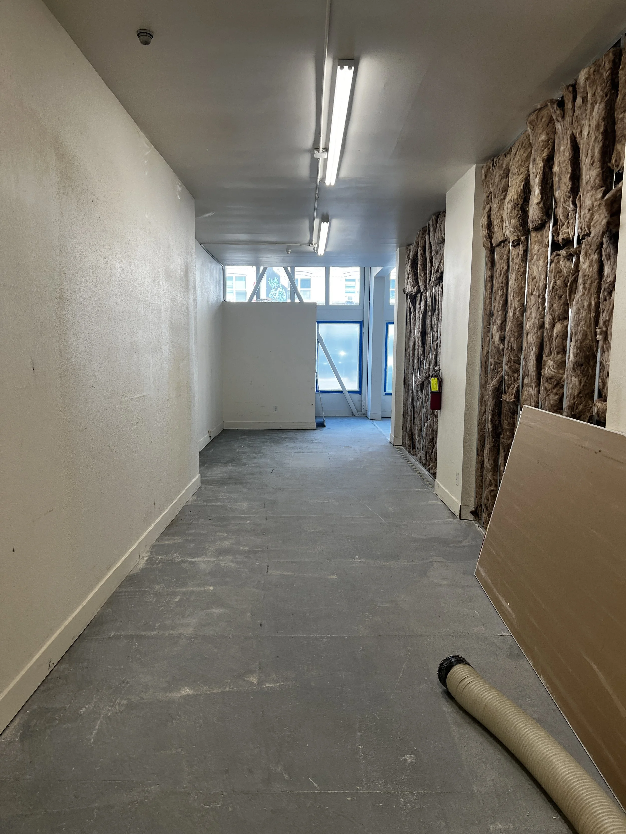 Interior of a building under construction with unfinished walls, exposed insulation, a large window at the end, and construction materials on the floor.