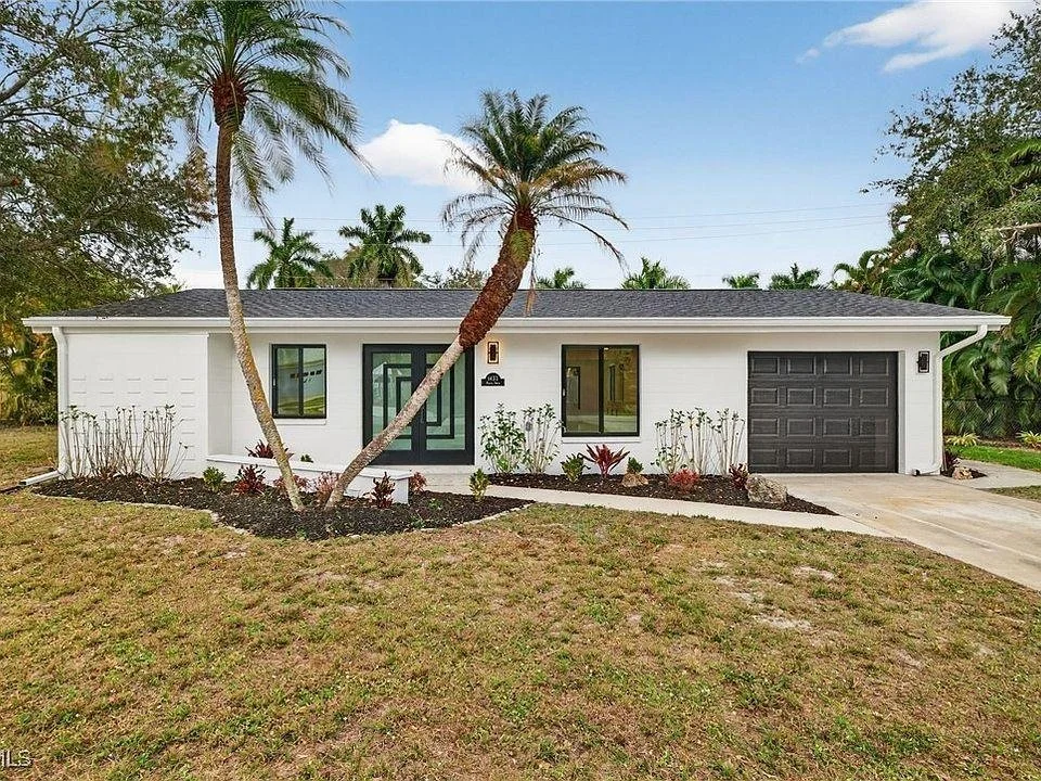 Front view of a modern, single-story house with a white exterior, black front door, large windows, and an attached garage. There are tall palm trees and landscaped bushes in the yard.