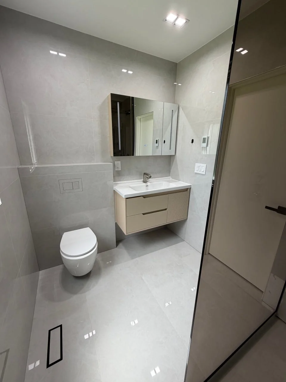 Modern beige bathroom with floating vanity, mirror cabinet, toilet, and reflective tiles.