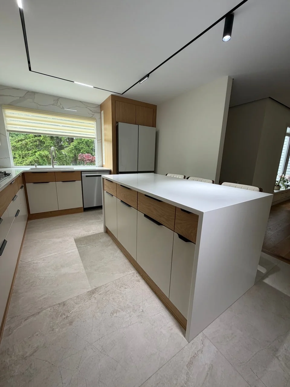 Modern kitchen with white and wood cabinets, a large white island, a window with a view of trees, and recessed ceiling lights.