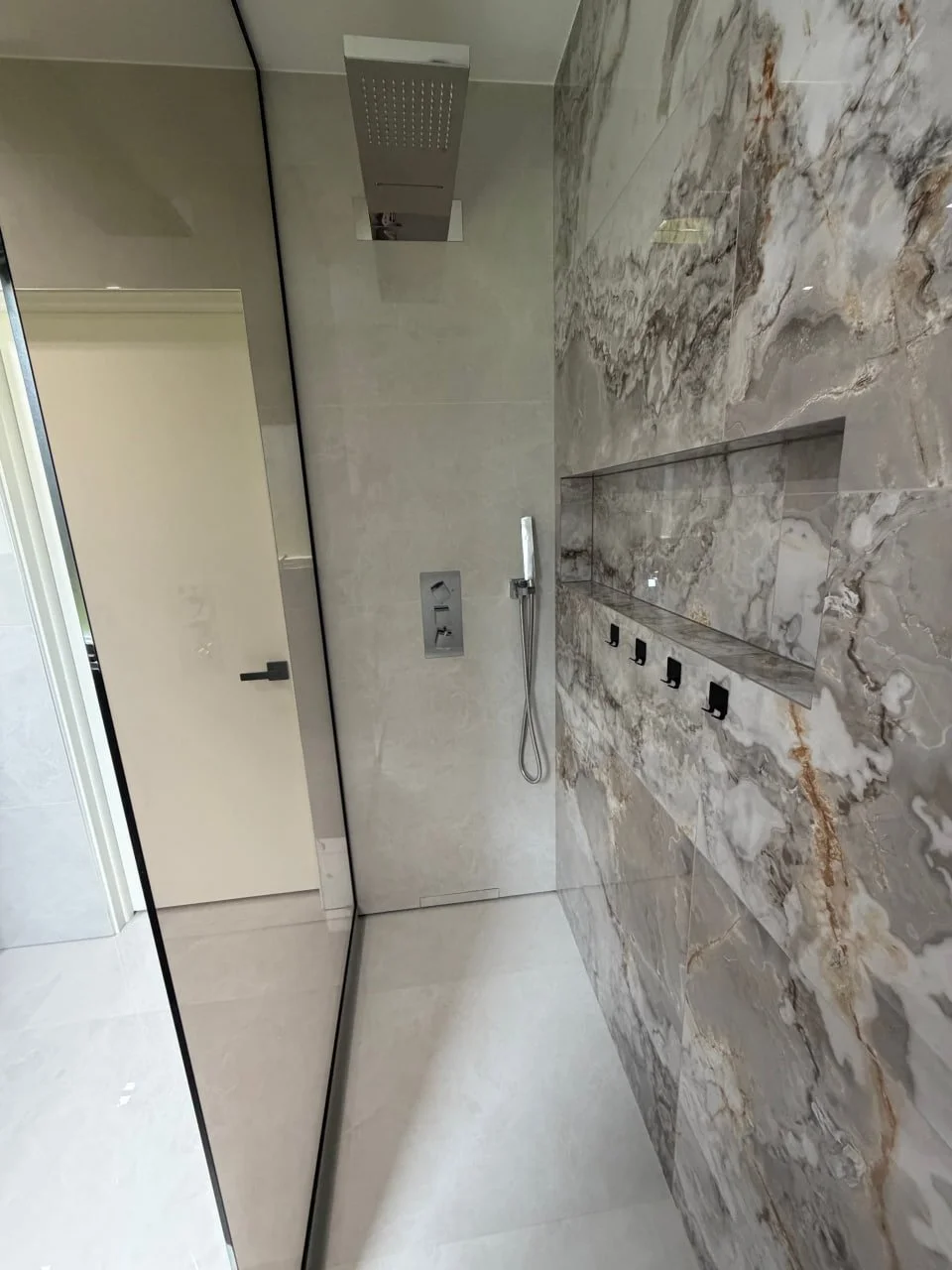 Modern bathroom shower area with marble wall tiles, a large rainfall showerhead, a handheld shower wand, and four black hooks along the marble wall.