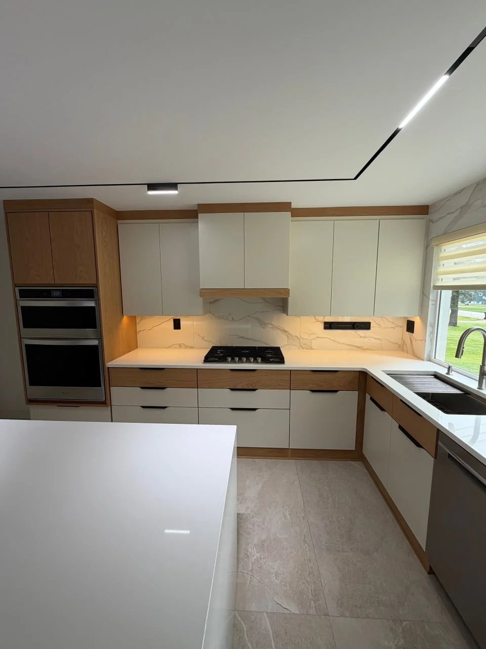Modern kitchen with white cabinets, wooden accents, marble backsplash, black stovetop, stainless steel appliances, and a window with a view of green outdoor space.