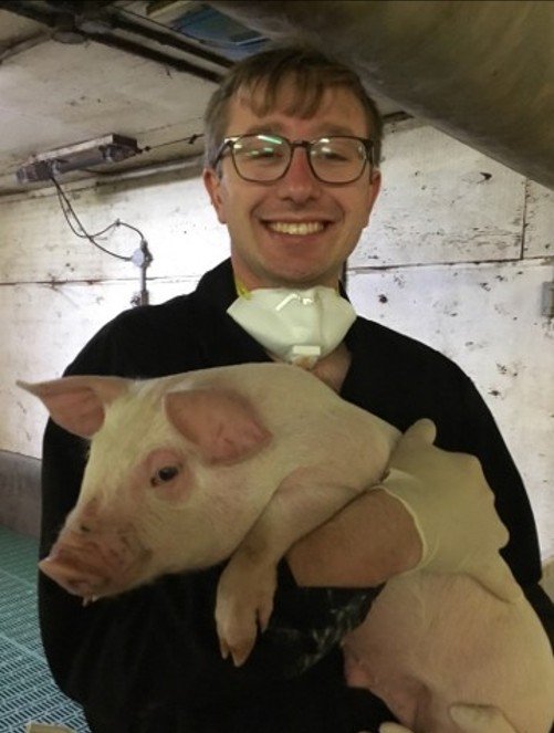 Smiling man wearing glasses holds a pink piglet in an indoor barn or utility room.