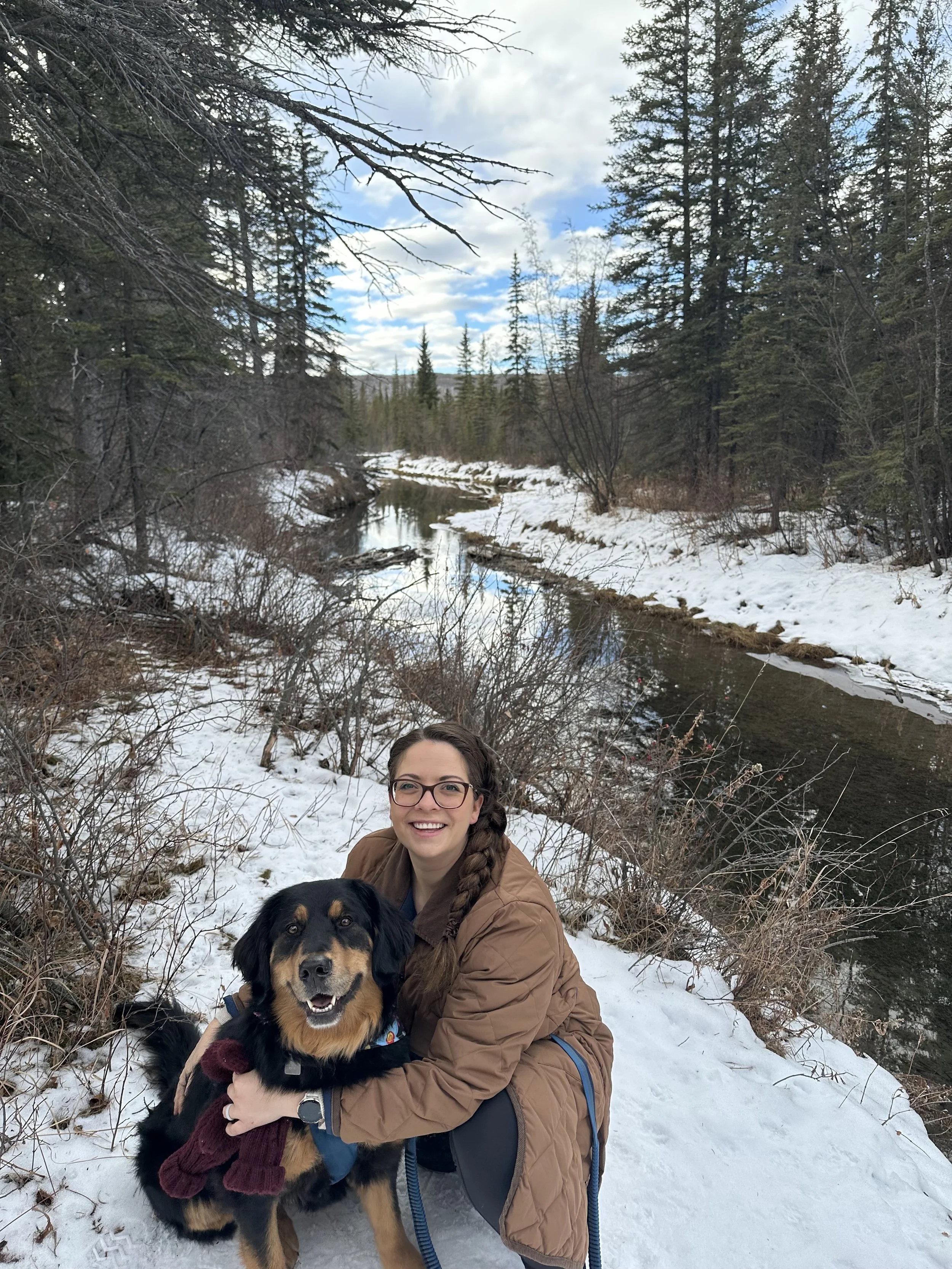 Woman hugging a large black-and-tan dog beside a partially frozen creek in a snowy evergreen forest.