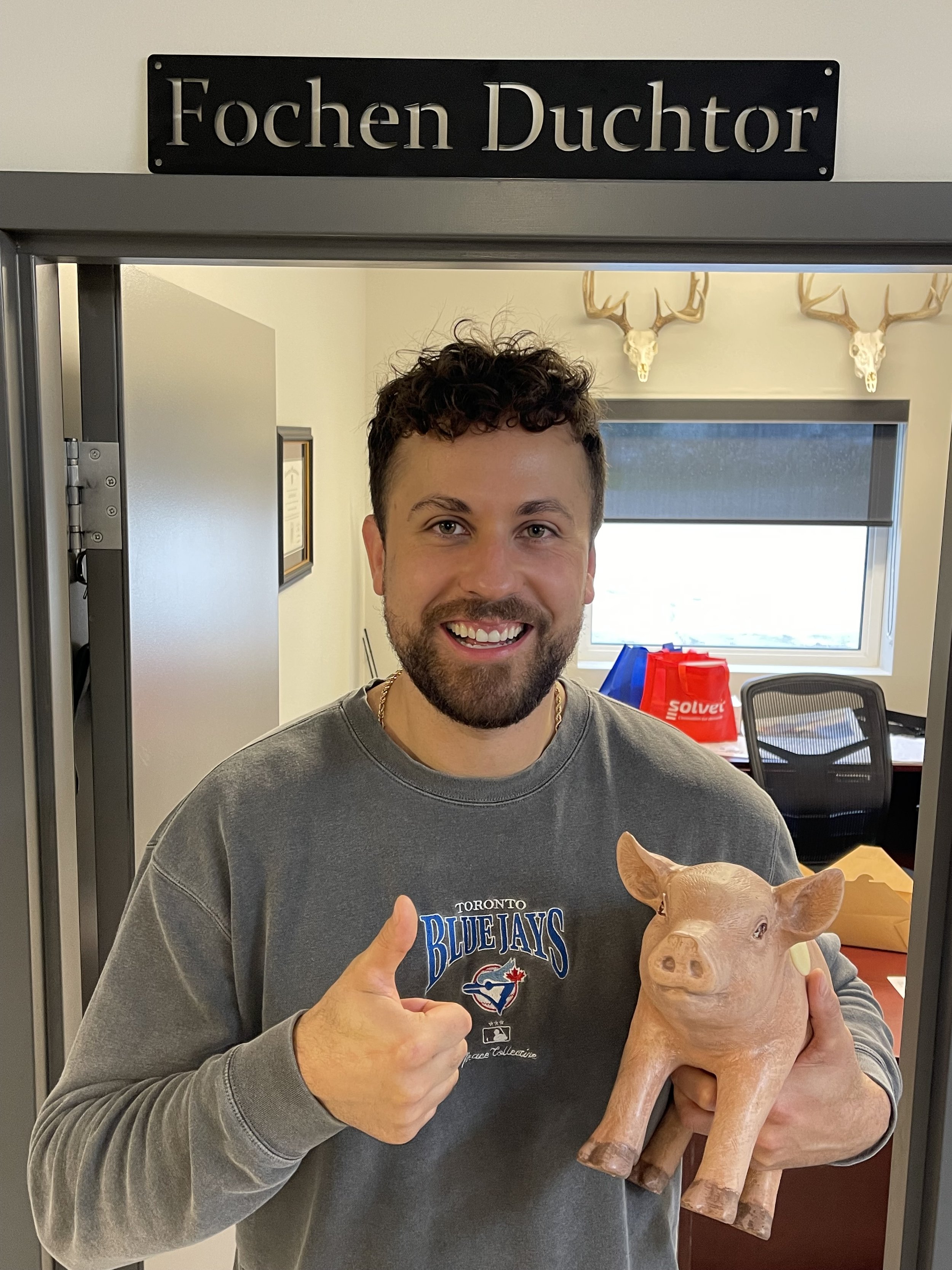 Smiling man in a Toronto Blue Jays sweatshirt holds a pig statue and gives a thumbs-up beneath a “Focken Realtor” sign.