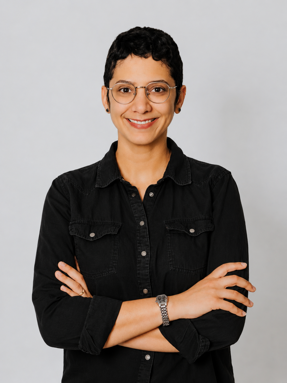 Smiling person wearing round glasses and a black button-up shirt, standing with arms crossed against a gray background