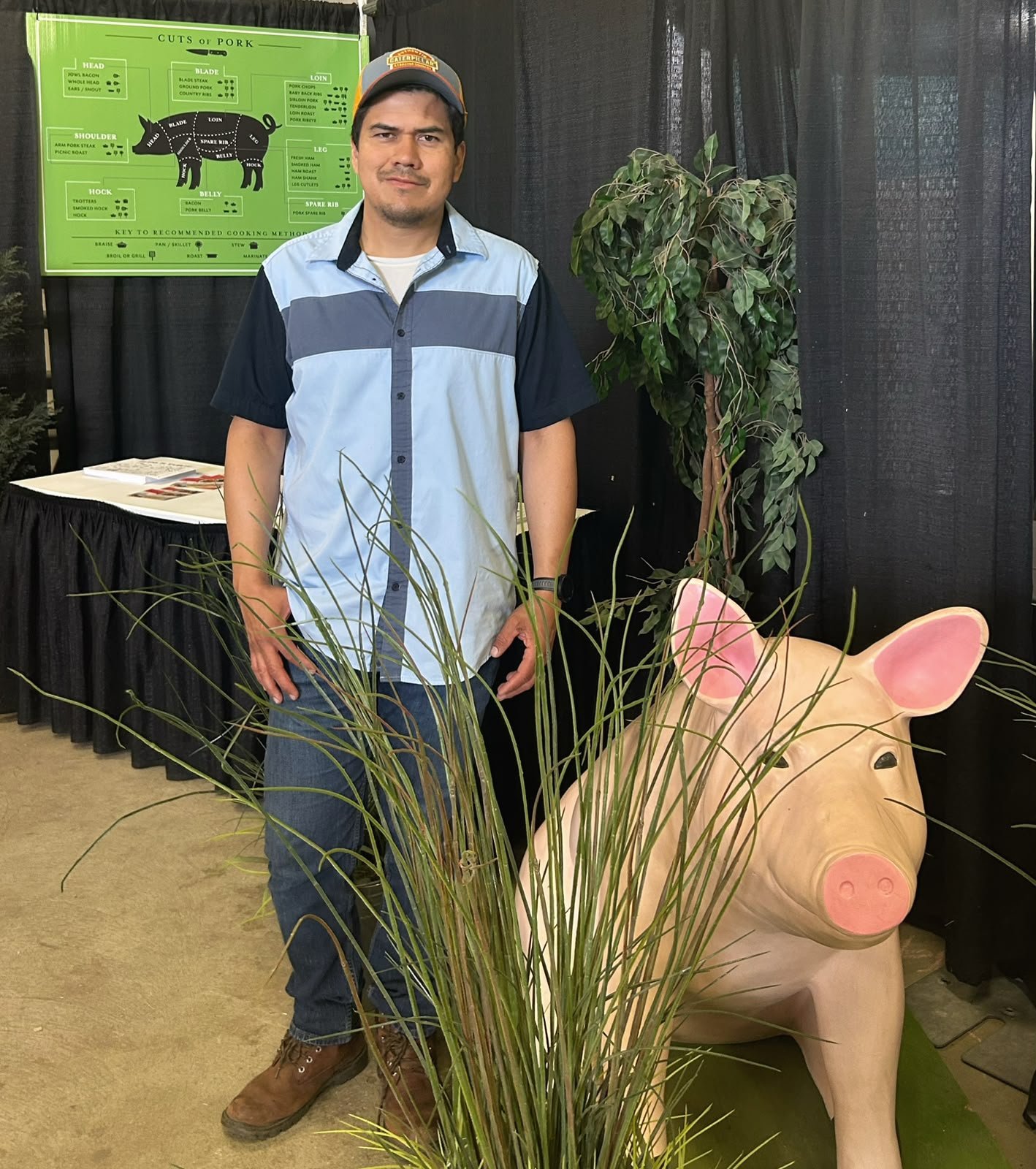 Man standing beside a large pig sculpture at an agricultural exhibit, with a cuts of pork chart and decorative plants.