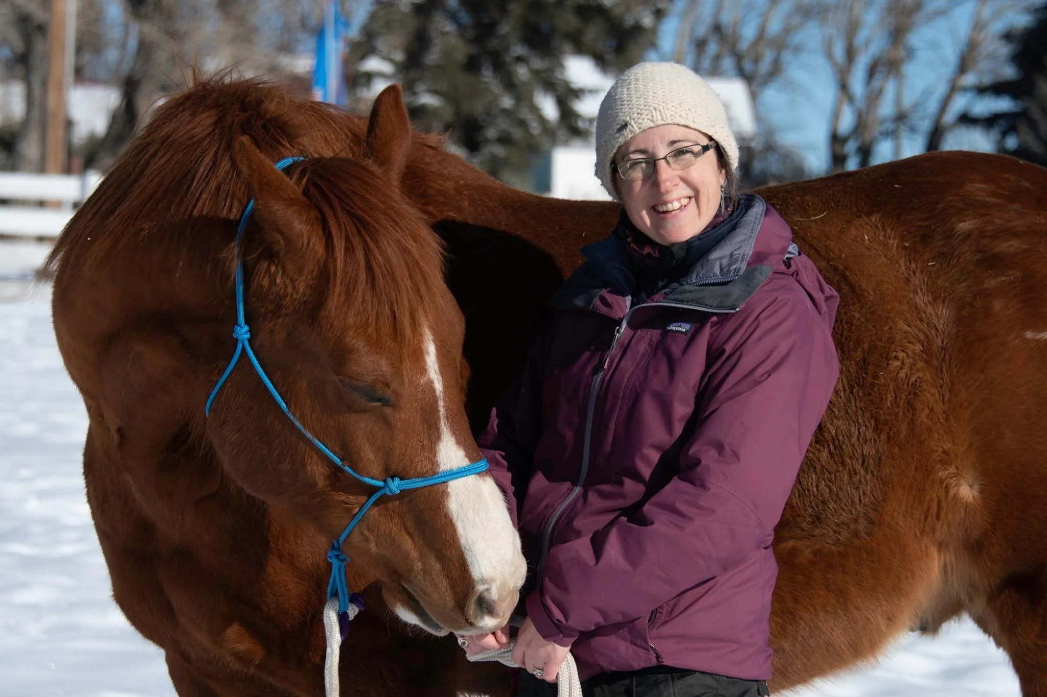 Smiling woman in winter clothing standing beside a chestnut horse in a snowy outdoor setting