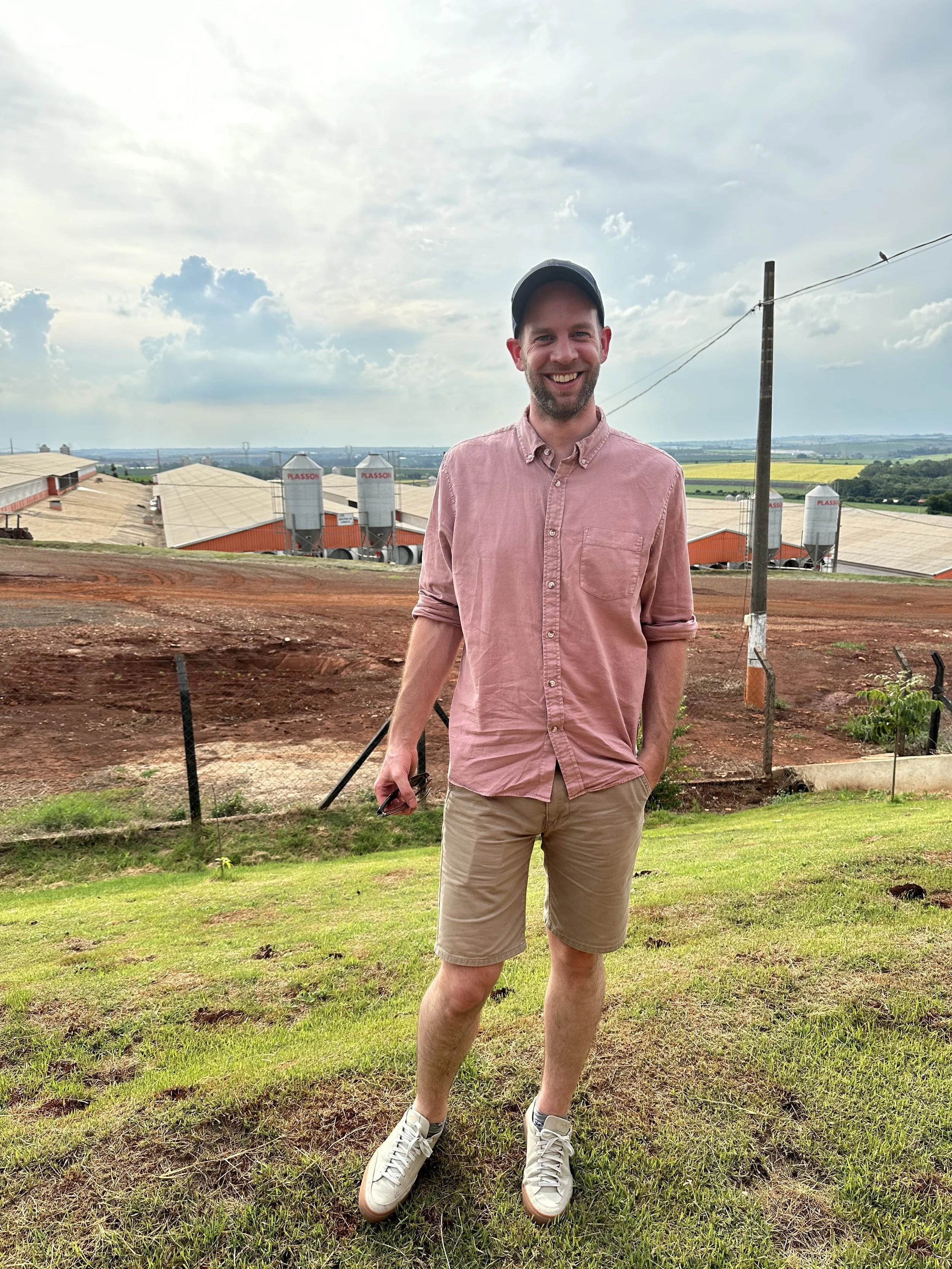 Smiling man in a pink shirt and cap standing on a farm overlooking agricultural fields and grain silos.