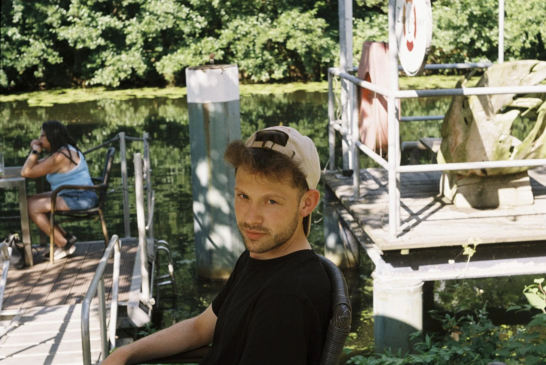 Junger Mann mit Baseballkappe sitzt im Freien, im Hintergrund See, grüne Bäume, Frau am Wasser sitzend, Brücke, Wasserpflanzen.
